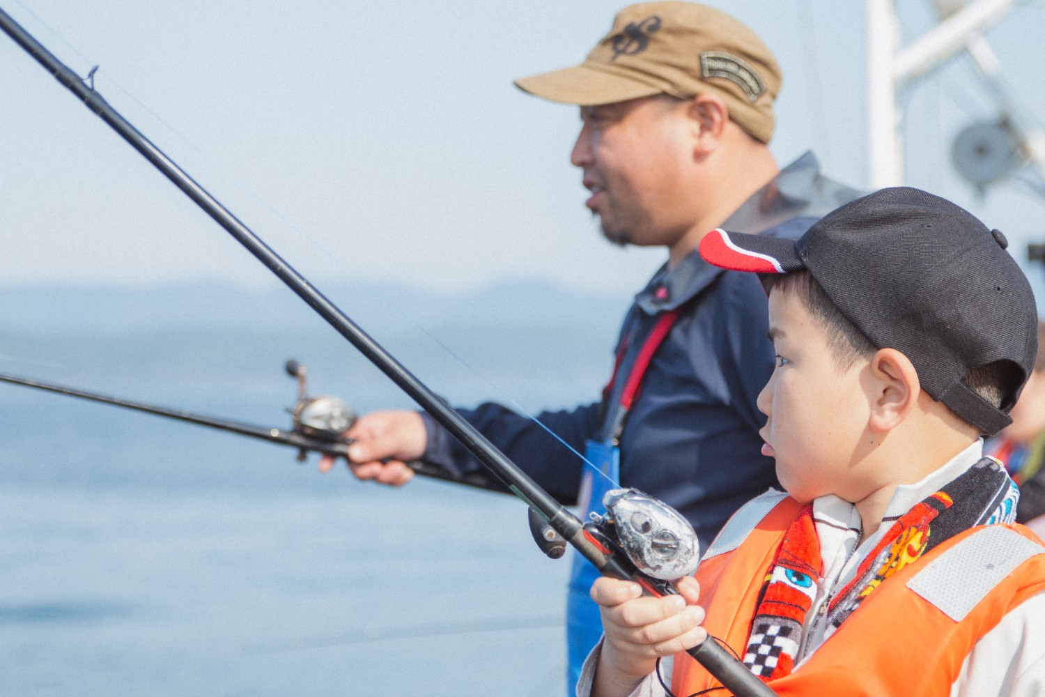 父親とライフジャケットと帽子を着けて真剣に釣り竿を握っている芳雅くんが並んで釣りをしている様子を横から撮影した写真
