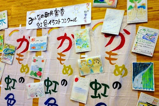 体育館の壁に貼られた「かもめの虹色談義」と書かれた横断幕の上に、子どもたちのカラフルな絵が多数展示されている写真