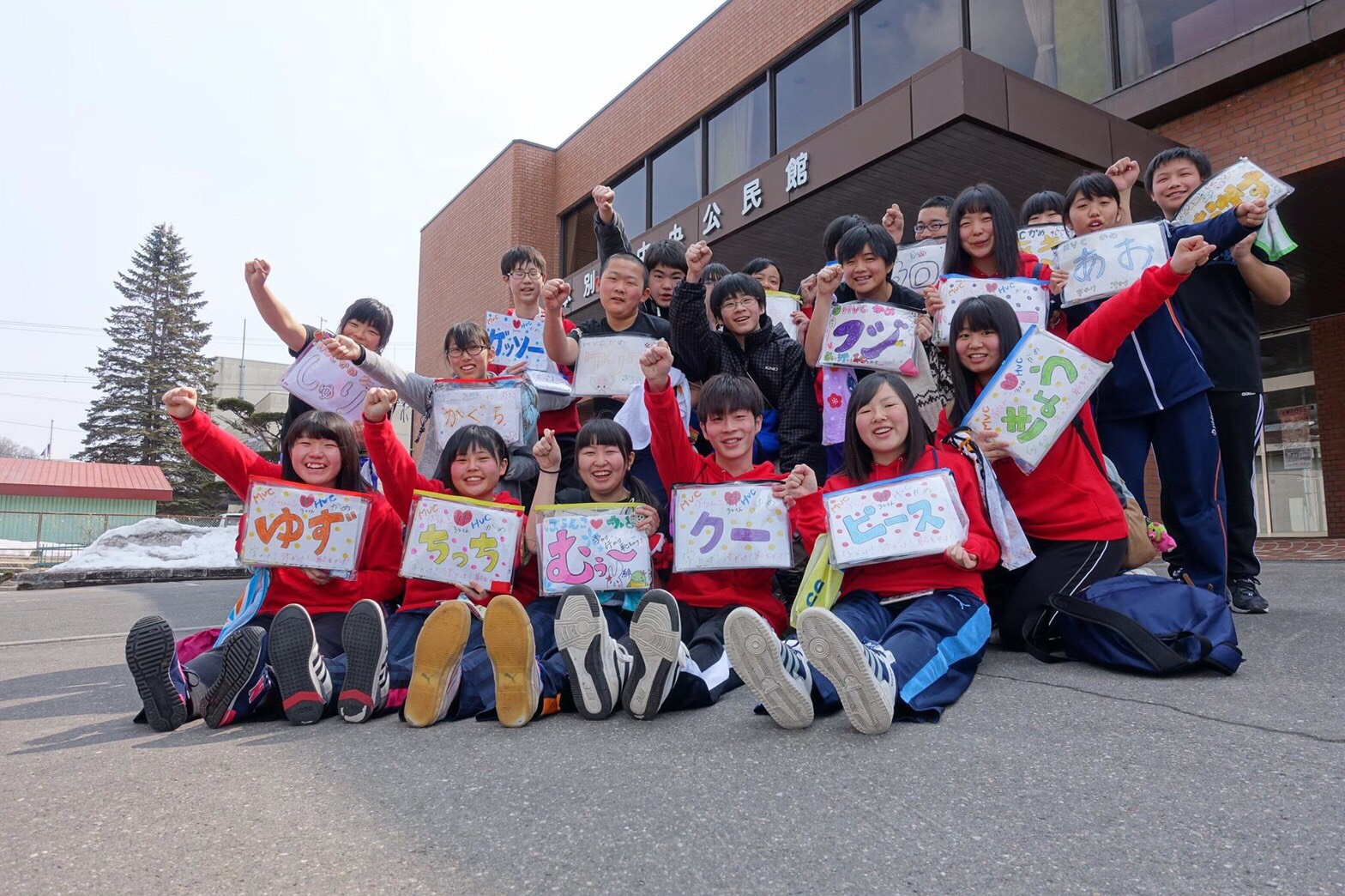 赤や青のジャージを着た十数人の高校生が、公民館の前で手書きのニックネーム入りのカラフルなプレートを持ち、腕を上げて元気にポーズを取りながら笑顔で集合写真に写っている様子の写真