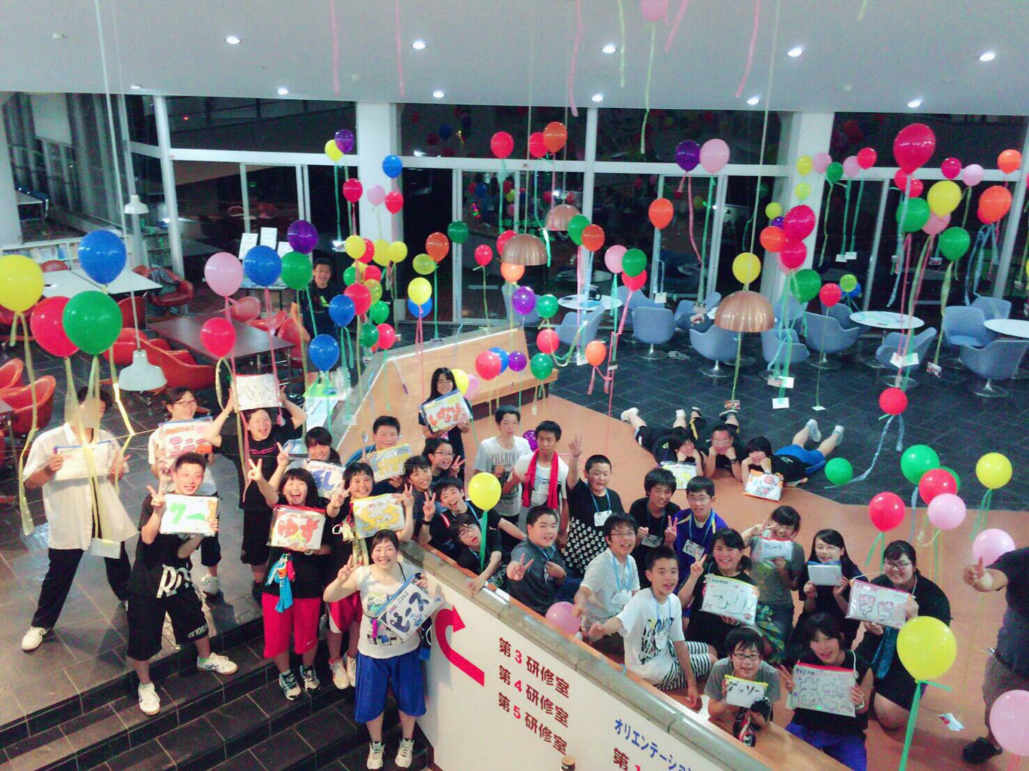 カラフルな風船が天井から吊るされた明るい室内で、名札を持った多くの若者たちが笑顔でピースをしながら集まって記念撮影をしている写真