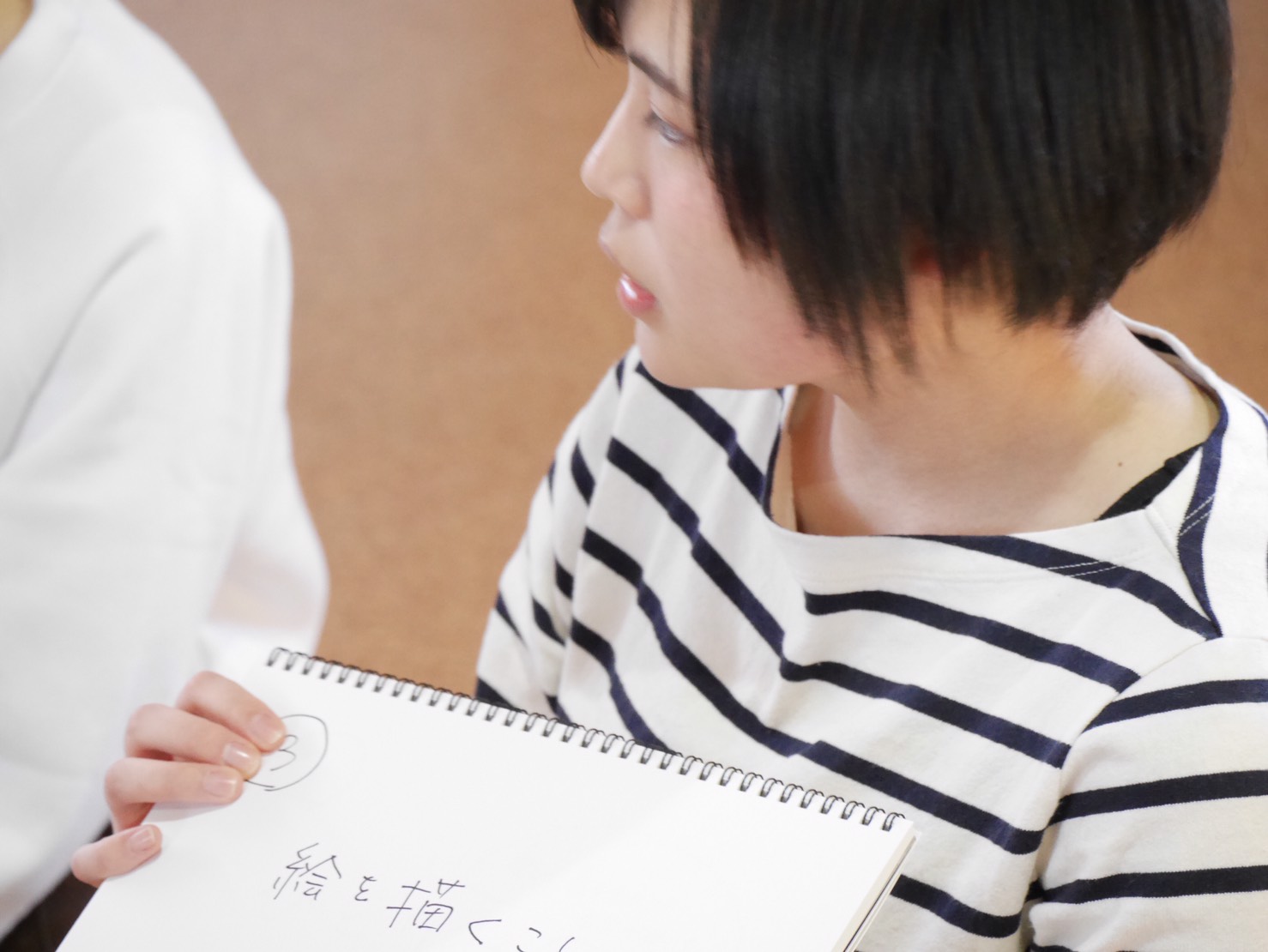 「絵を描くこと」とかかれたスケッチブックを手に持っているボーダーの服を着た三浦千裕さんの写真