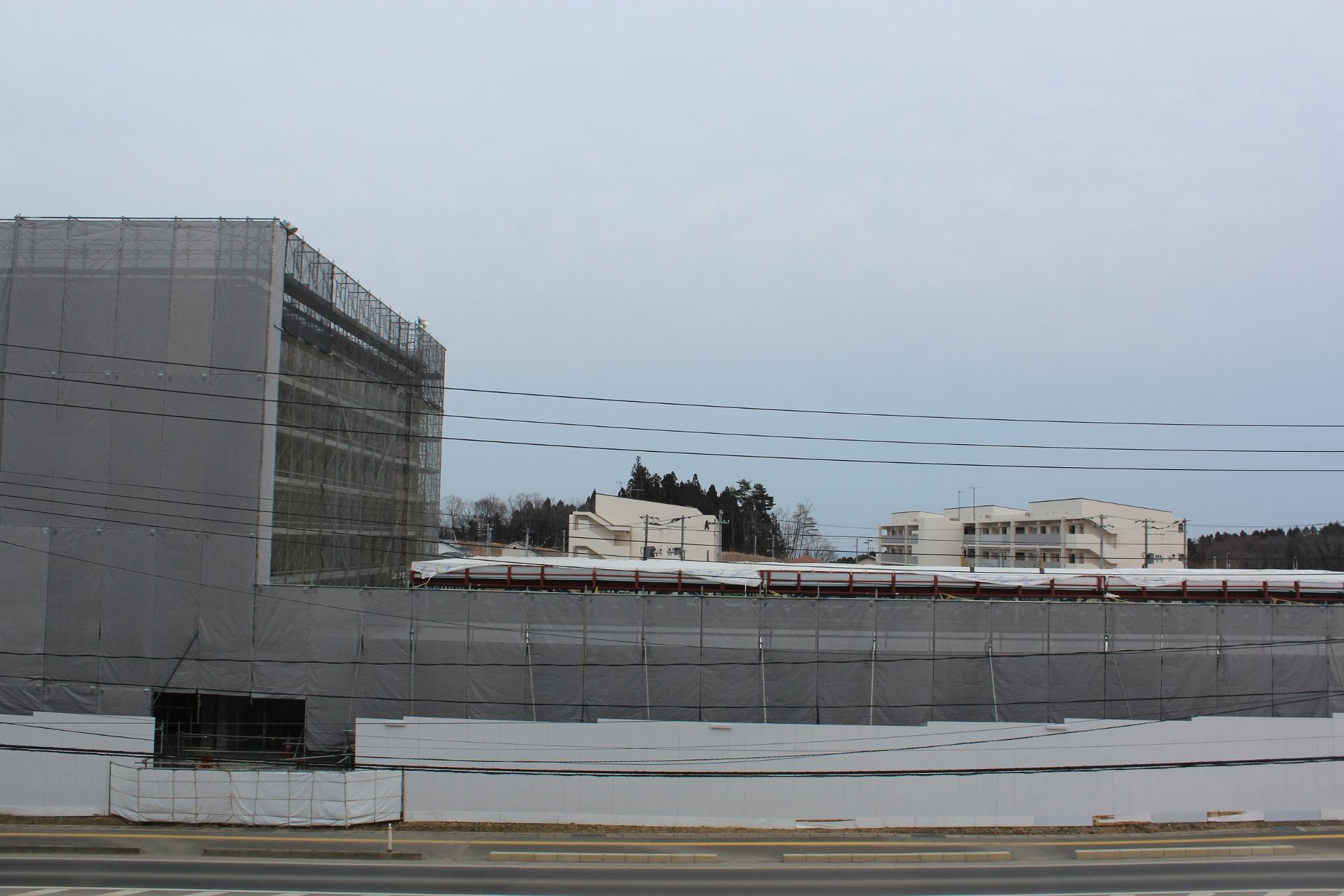 足場と防護ネットに覆われた建設中の建物が道路越しに見える平成29年3月6日現在の写真