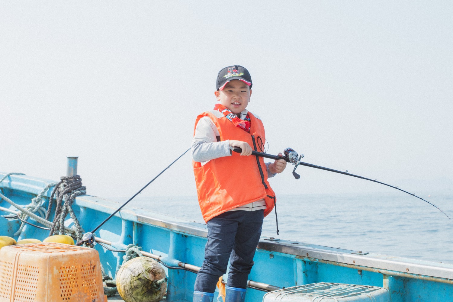 オレンジのライフジャケットを着た芳雅くんが青い船の上で釣り竿を持ち、笑顔でカメラに向かって立っている姿を写した写真