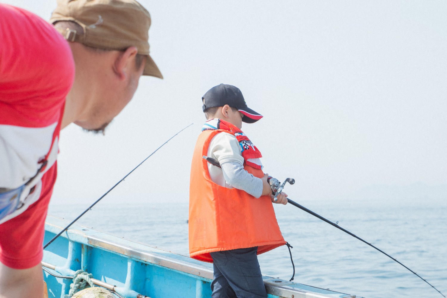 オレンジ色のライフジャケットと黒い帽子を着用し釣りをしている芳雅くんと、赤いTシャツとベージュの帽子をかぶった父親が青い船の上で海に向かって釣りをしている様子の写真
