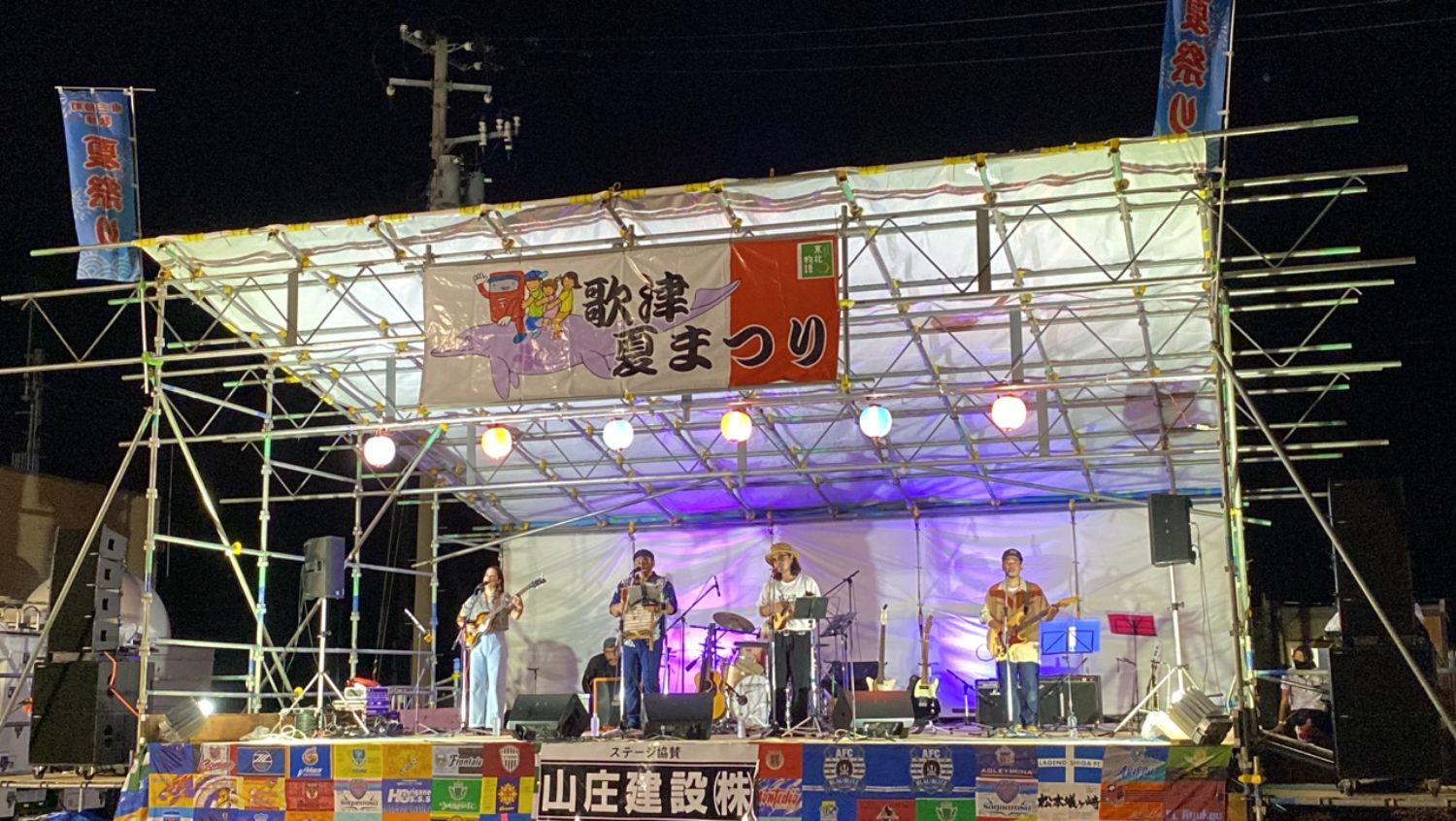 「歌津夏まつり」と書かれた夜のステージでバンドの演奏者が演奏している様子の写真