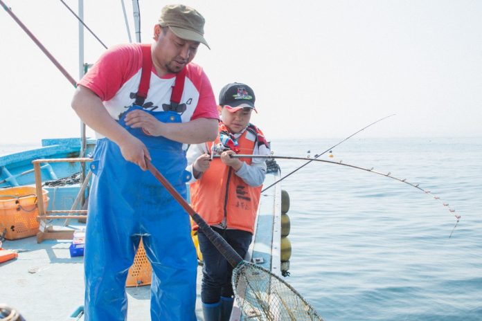 青いオーバーオールとベージュの帽子をかぶりながら釣り竿を持つ父親と、隣にいる黒い帽子とオレンジのライフジャケットを着た芳雅くんが一緒に釣りをしている様子を船の上で写した写真