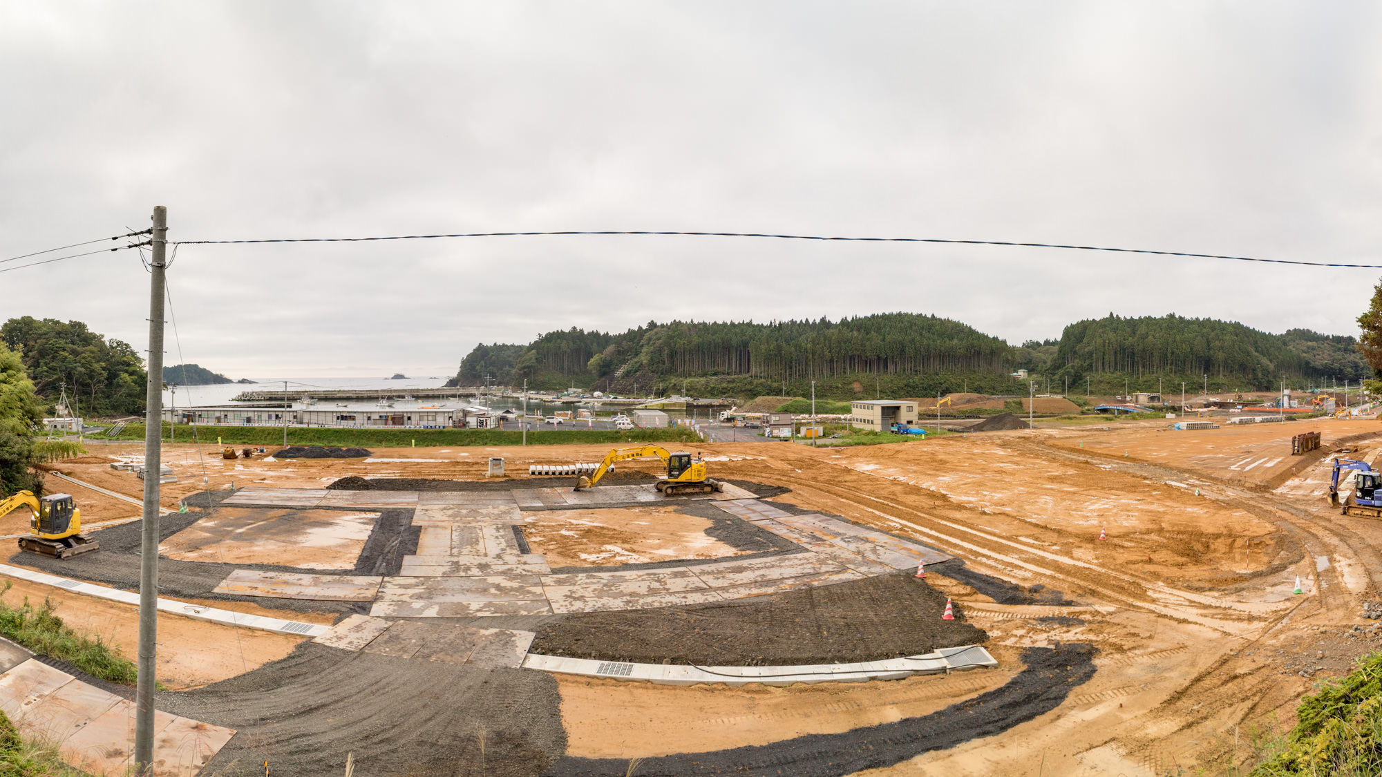 複数のショベルカーが稼働している建設現場で、地面には新しく舗装されたアスファルトや土が見られ、工事が進行されており背景には建物と木々に覆われた森林地帯が広がっている写真