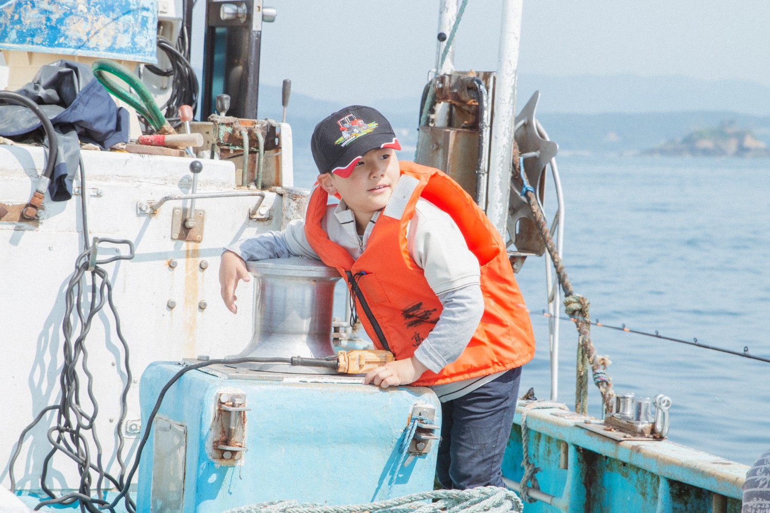 オレンジ色のライフジャケットと黒い帽子を着用した芳雅くんが、青い船の上でタンクの様な物に手をかけて海を見つめている写真