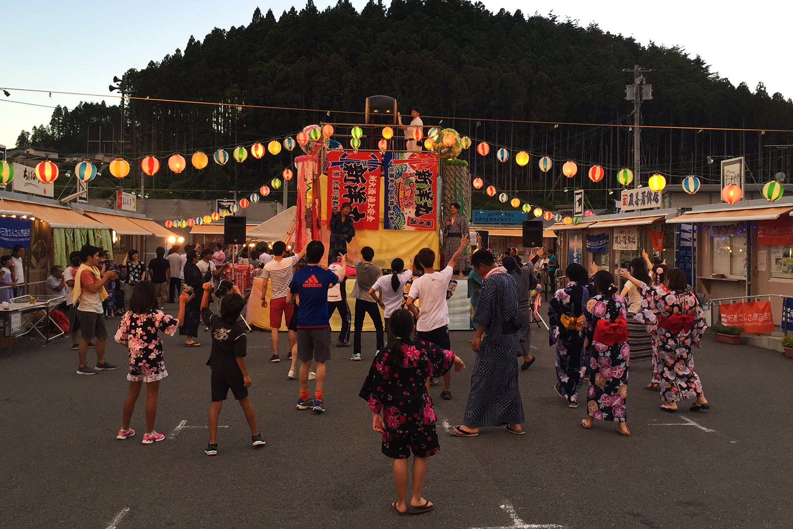 夕暮れ時の空の下、色とりどりの提灯が灯る中、中央のやぐらを囲んで浴衣姿の人々が楽しそうに盆踊りを踊っている、にぎやかで温かい雰囲気の夏祭りの様子の写真