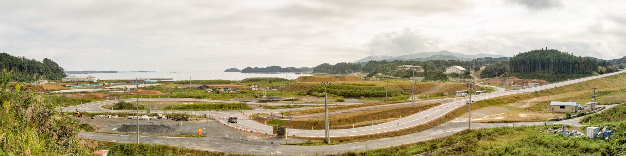 手前には曲がりくねった道路があり、その周囲にはいくつかの建設現場や建物が点在し背景には木々や草に覆われた丘が広がり、画像の左側には海が見える海沿いの地域を広く見渡したパノラマ風景の写真