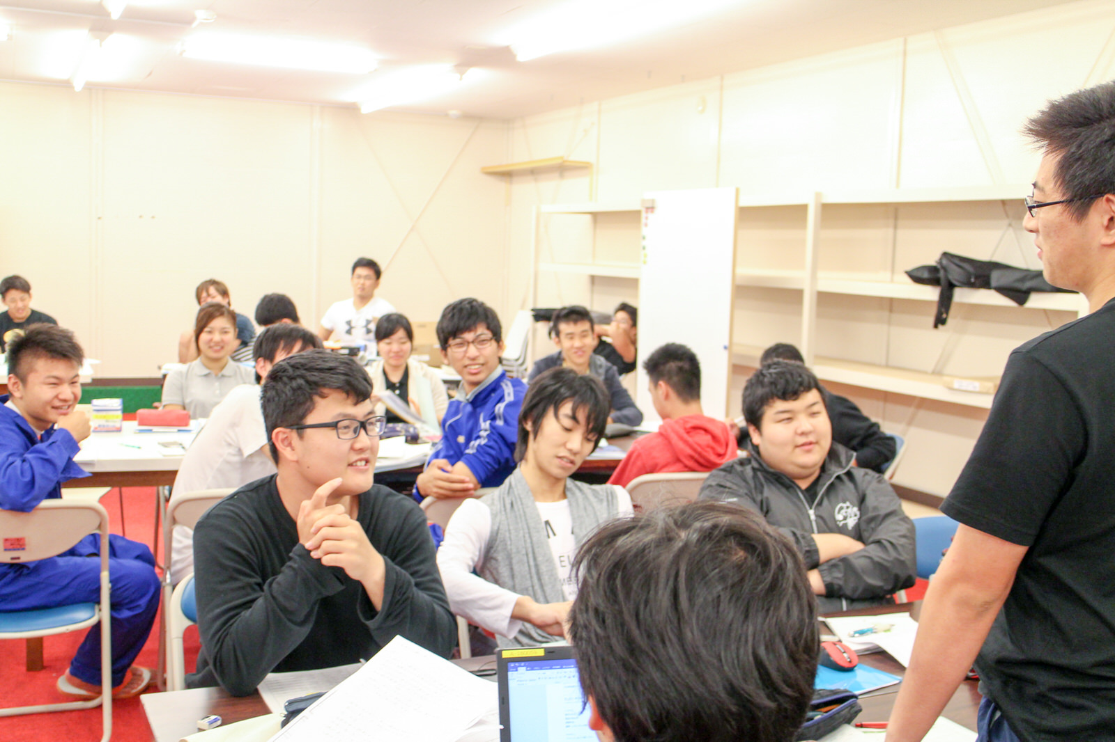 教室で前方に立っている男性の話を、資料が置かれているテーブルの席に座った多数の若者たちが和やかに聞いている写真