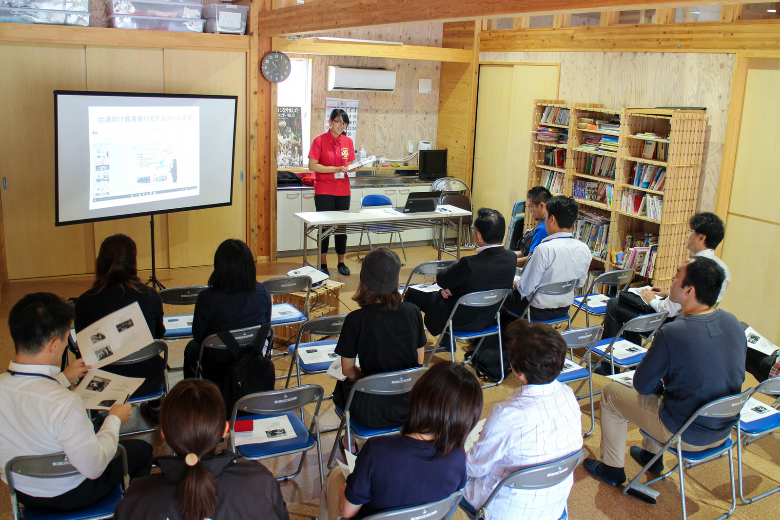 本棚が置かれた木の壁の室内で、並んだパイプ椅子に座った参加者たちが、資料が映し出された前方のスクリーンと傍に立つ赤いポロシャツの女性の話を、資料を手に持ちながら聞いている写真