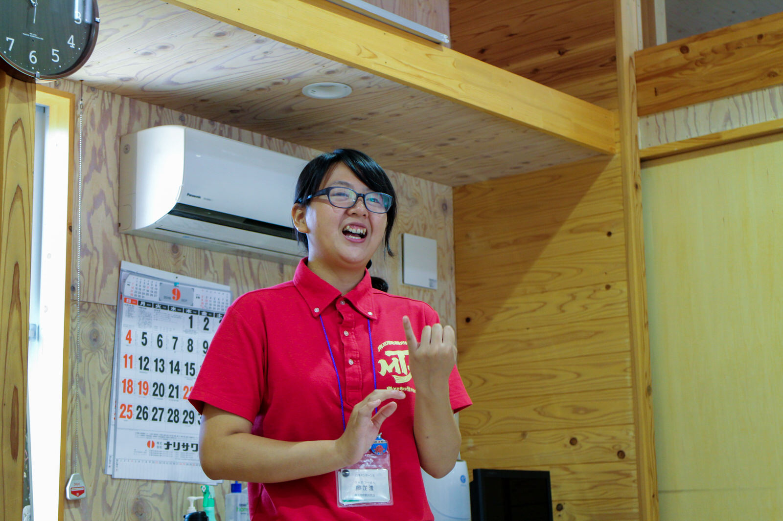 木の壁の室内で、赤いポロシャツを着て首から名札を下げた女性が立って指を折るジェスチャーをしながら話をしている写真