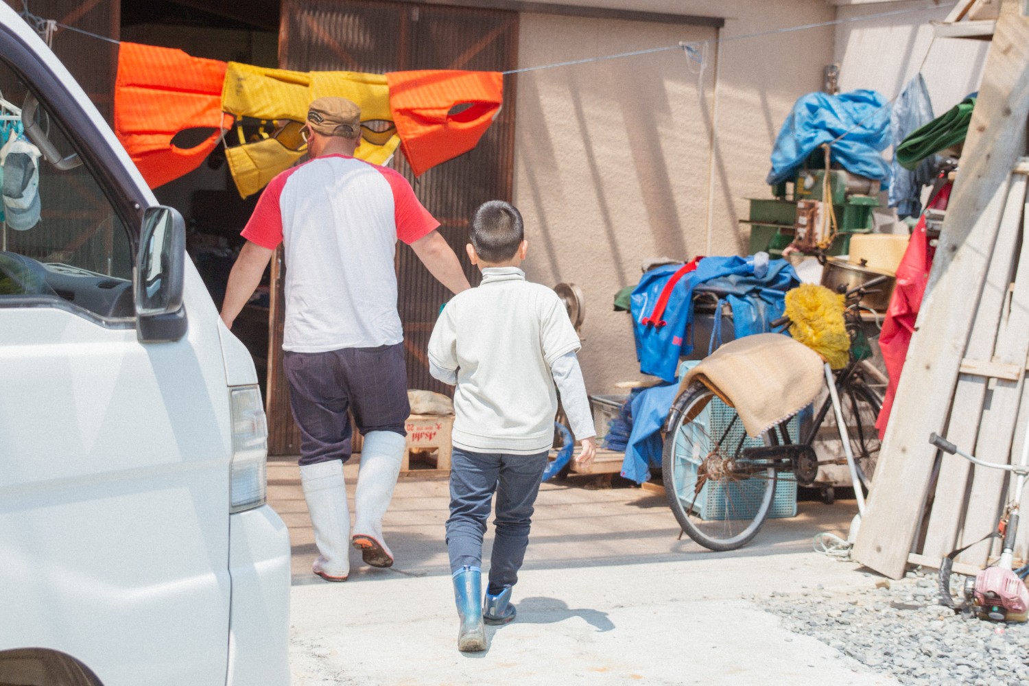 赤いTシャツを着た父親と、白い長袖シャツに青い長靴を履いた芳雅くんが、白い車の隣を歩いて古い自転車やカバーがかかった荷物のある建物の入口に向かって歩いている後ろ姿の写真