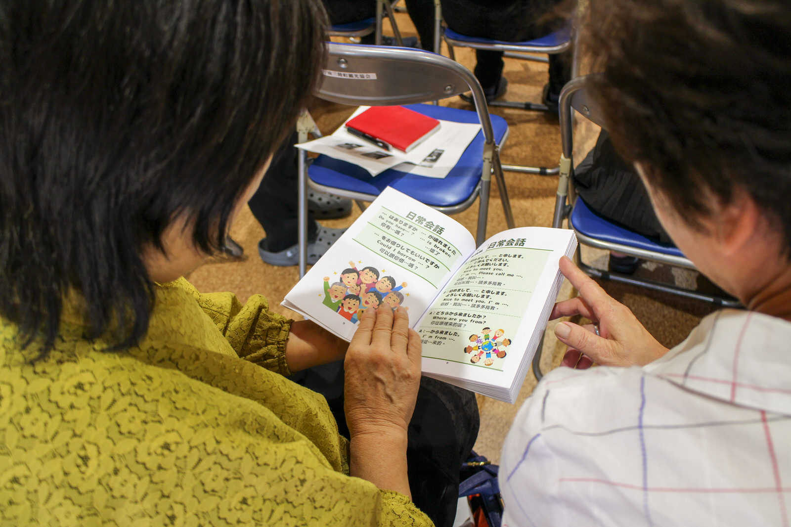 隣同士に座った2人の女性が、一冊の冊子を一緒に開き、イラスト付きのページを読んでいる様子を後方から撮影した写真