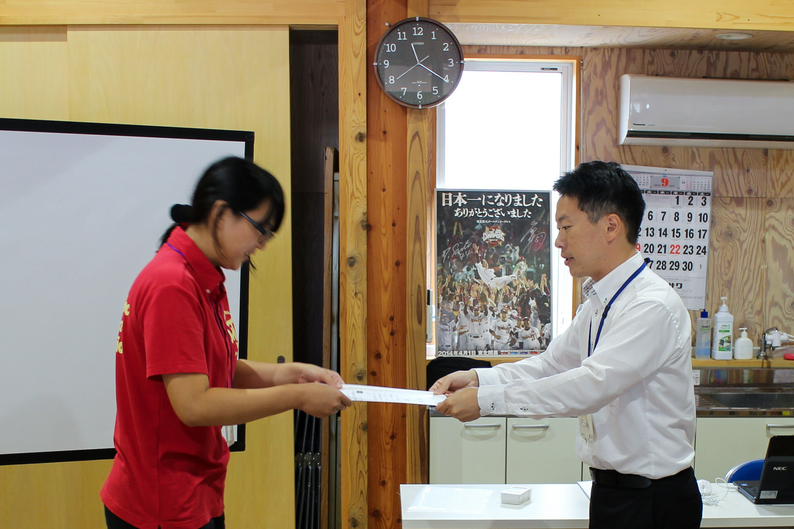 ポスターや壁掛け時計、カレンダーが飾られた室内で、赤いポロシャツを着た女性が、白いシャツと黒いズボンを着た男性から任命書を手渡されている様子の写真
