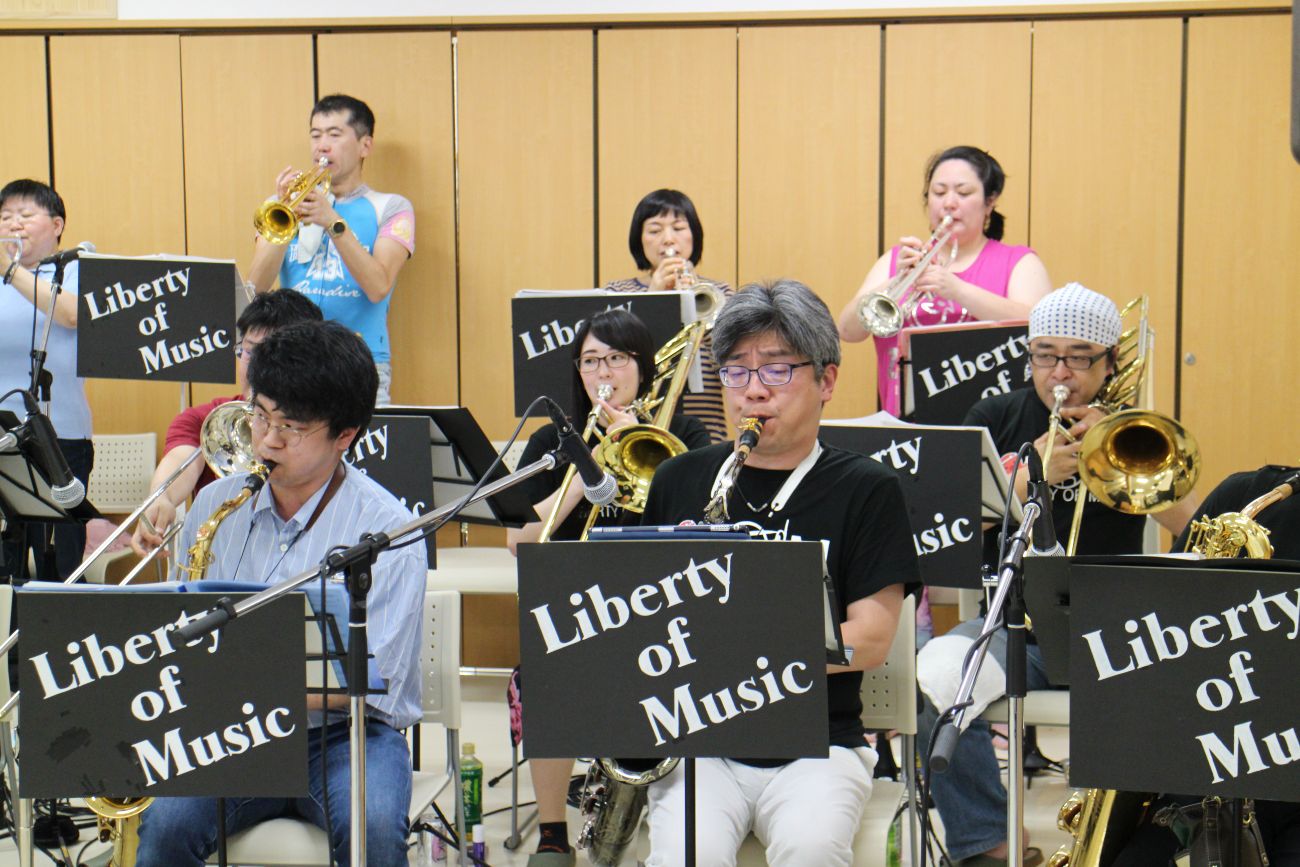 「Liberty of Music」と書かれた譜面台を前にして、メンバーがサックスやトランペット、トロンボーンなどの楽器を演奏している練習中の写真