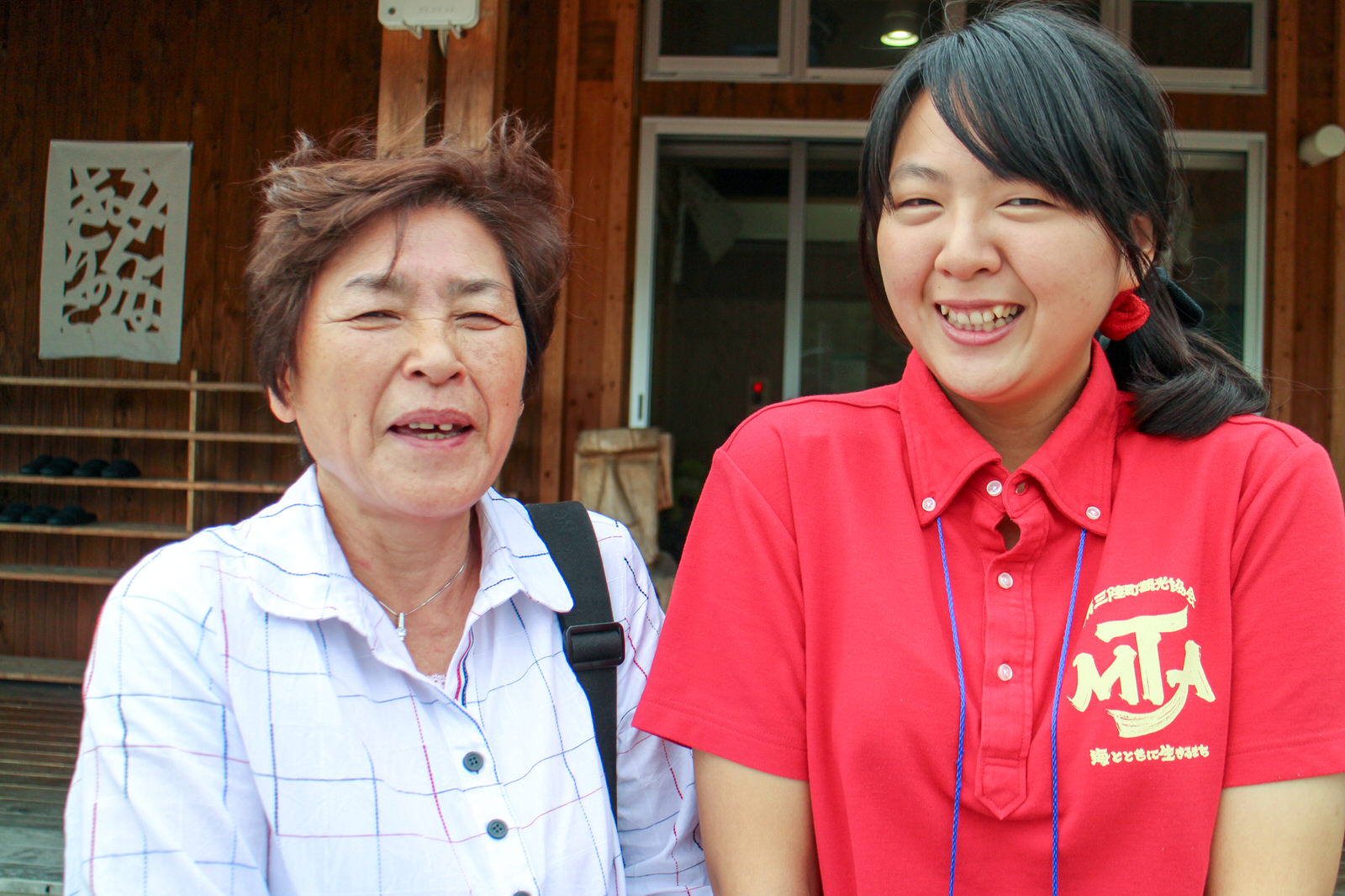 木造の建物の前で、白いチェック柄のシャツを着た女性と、赤いポロシャツを着ている女性が並んでいる記念写真