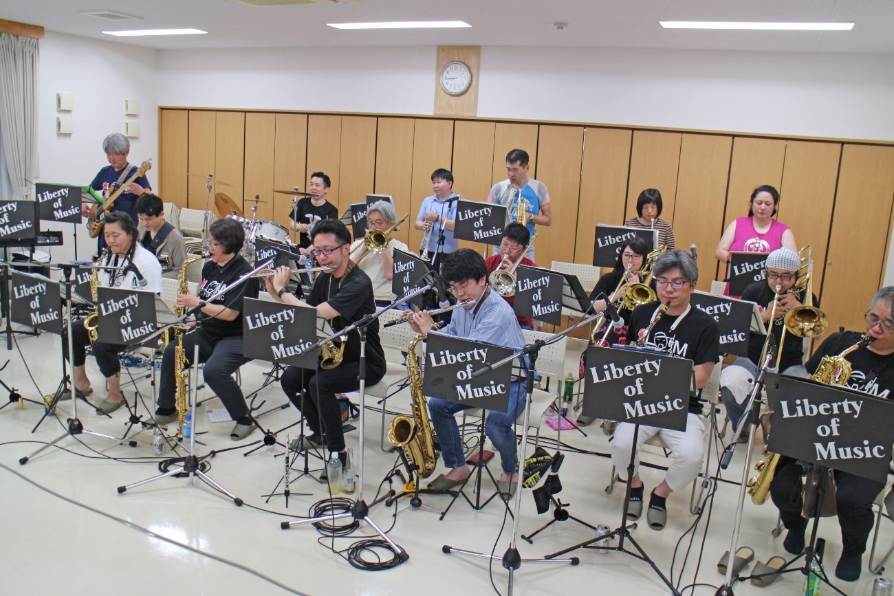 サックスやトロンボーン、トランペットにエレキギターを演奏するメンバーが、「Liberty of Music」の譜面台を前に音を合わせているリハーサル風景の写真