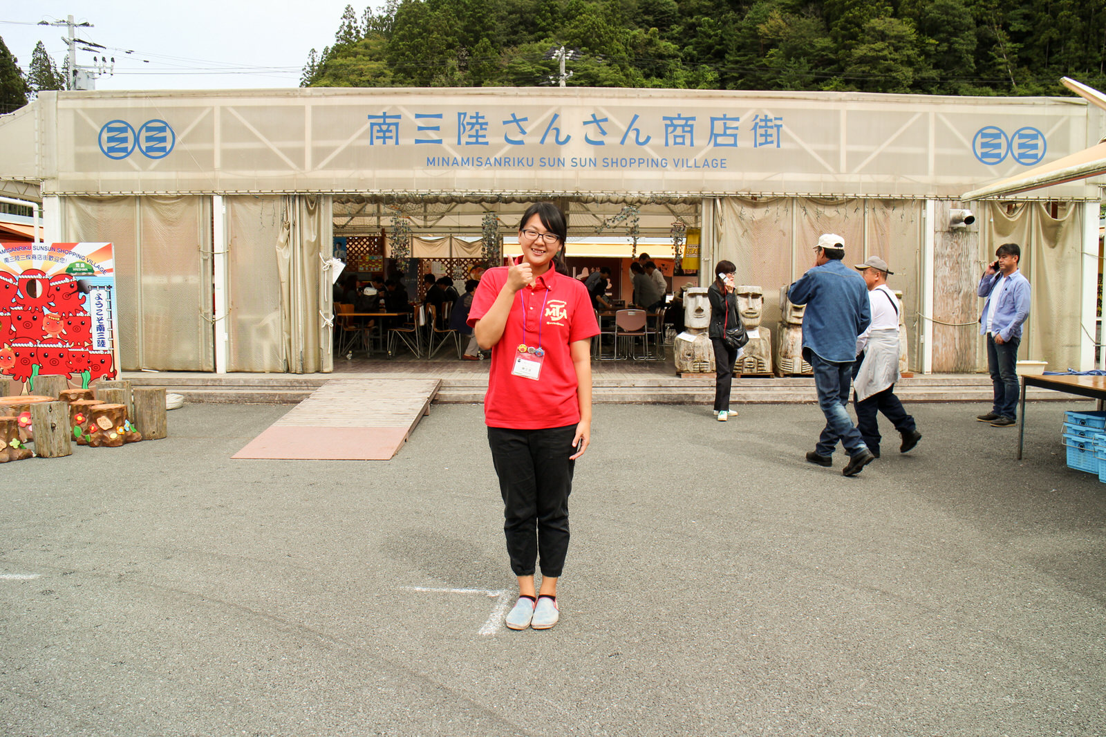 入口の左側にオクトパス君の顔出しパネルが置かれた「南三陸さんさん商店街」と書かれた建物の前に、赤いポロシャツを着た女性が立っている写真