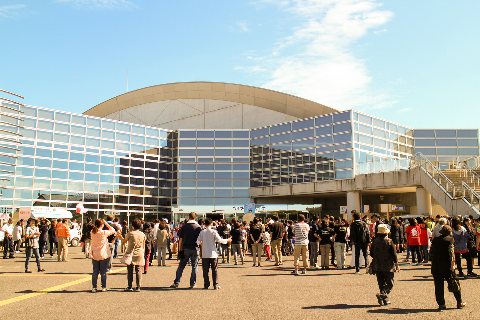 現代的なガラス張りの大きな建物の前に、様々な年代の人々が集まり、イベントの開始を待っている様子を晴天の下で撮影した写真