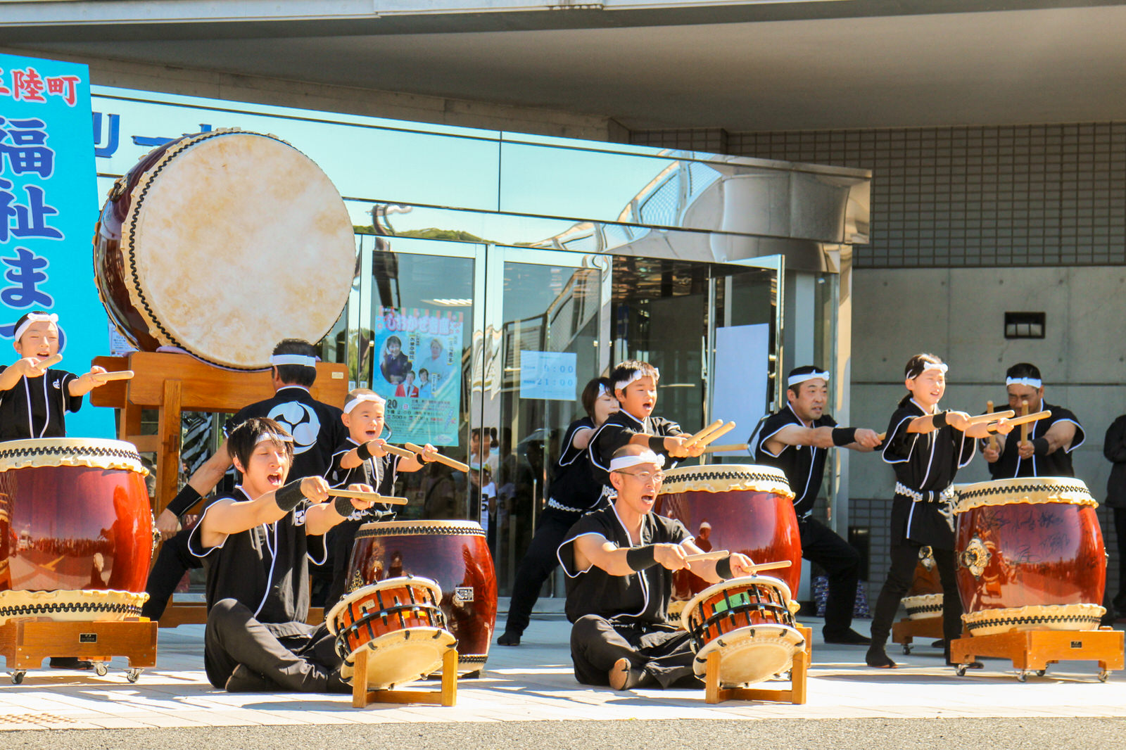 建物の入口前で黒い衣装を着た子どもから大人までの和太鼓グループ全員がバチを持った腕を前にさしながら力強く演奏している写真