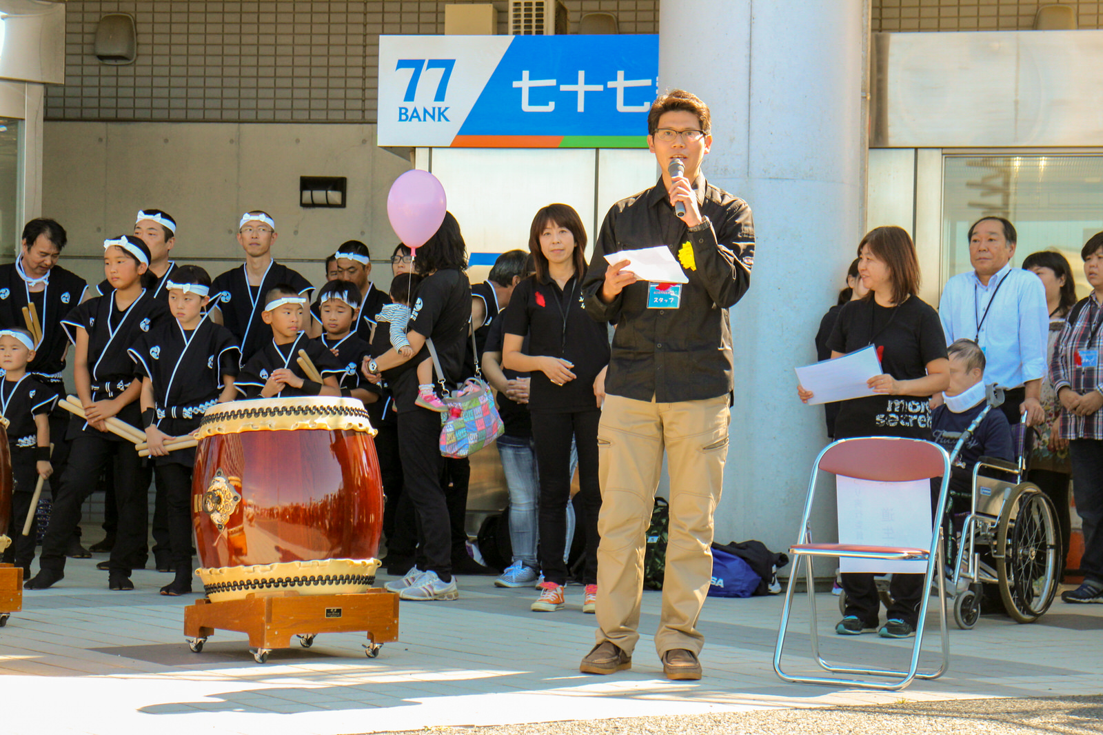 建物の屋外に設置された「77銀行」のATMの前に太鼓が並び、後方にお揃いの黒い衣装を着た子どもたちとそれを見守る大人たちが整列している傍で、黒い服を着た男性がマイクを持って挨拶を行っている様子の写真