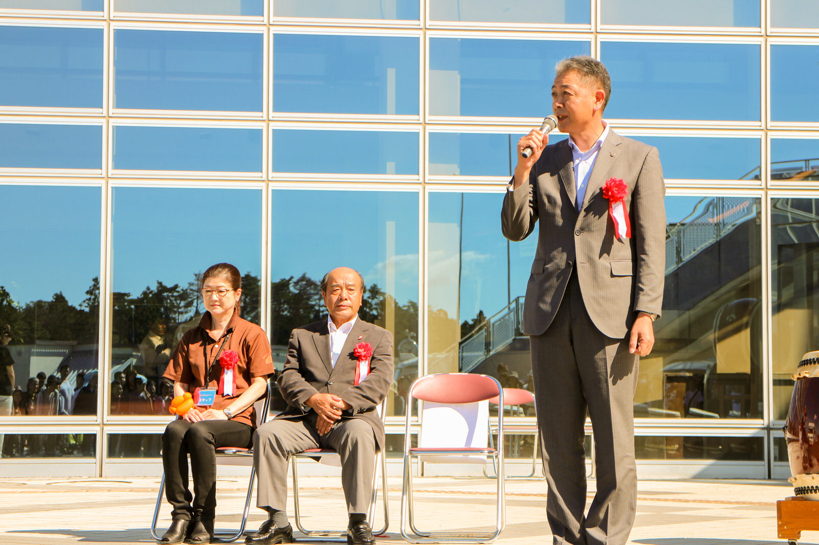 建物の前で椅子に座った二人の来賓と、スーツ姿で胸章リボンをつけマイクを持って話している副町長が立ち、開会式でのスピーチの様子を映した式典の写真