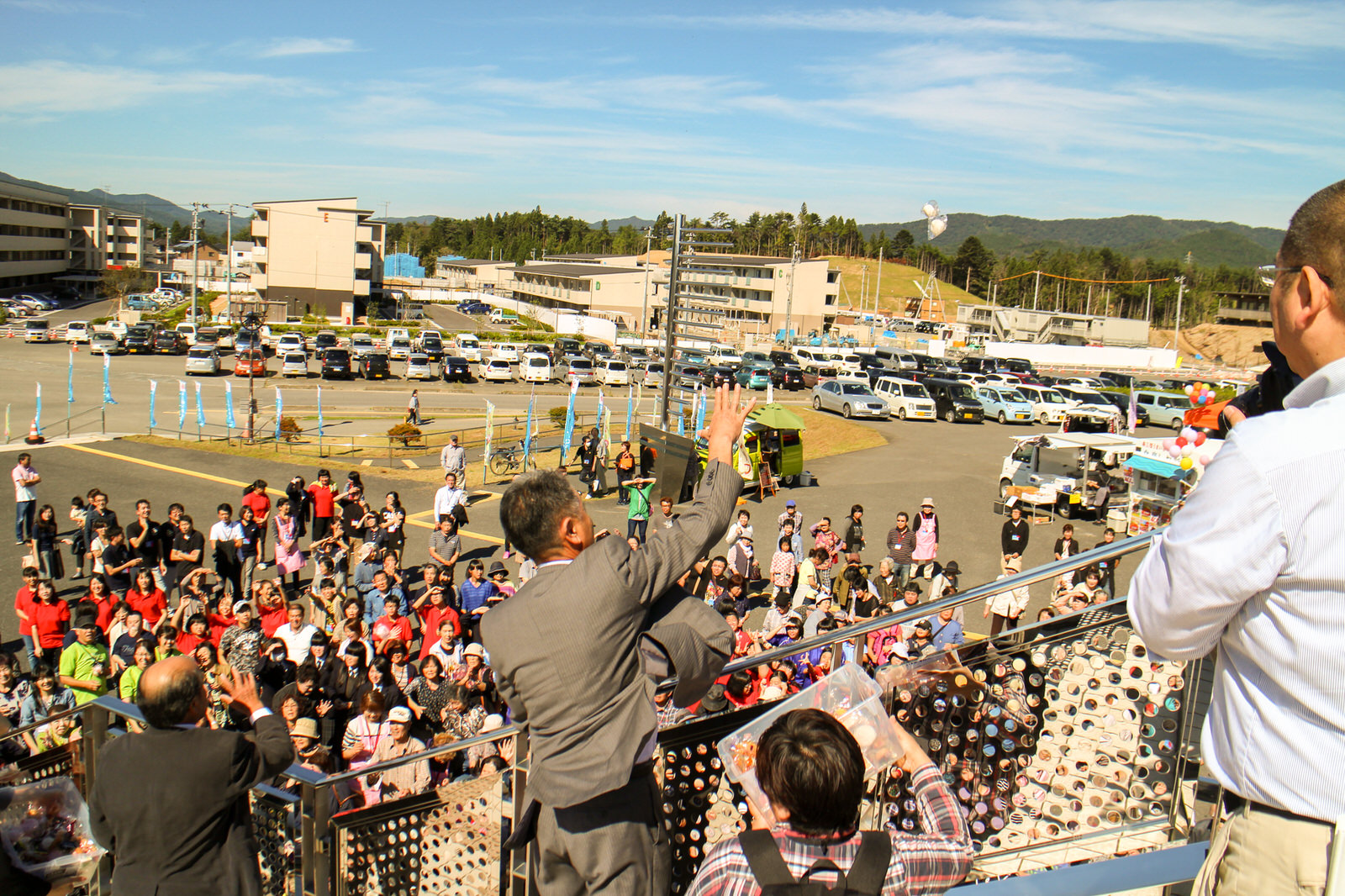 同じ階段の上から、来賓者やスーツ姿の副町長が片手を高く上げて群衆に餅を投げており、多くの人が手を伸ばしてそれを受け取ろうとしている賑やかな餅まきの写真