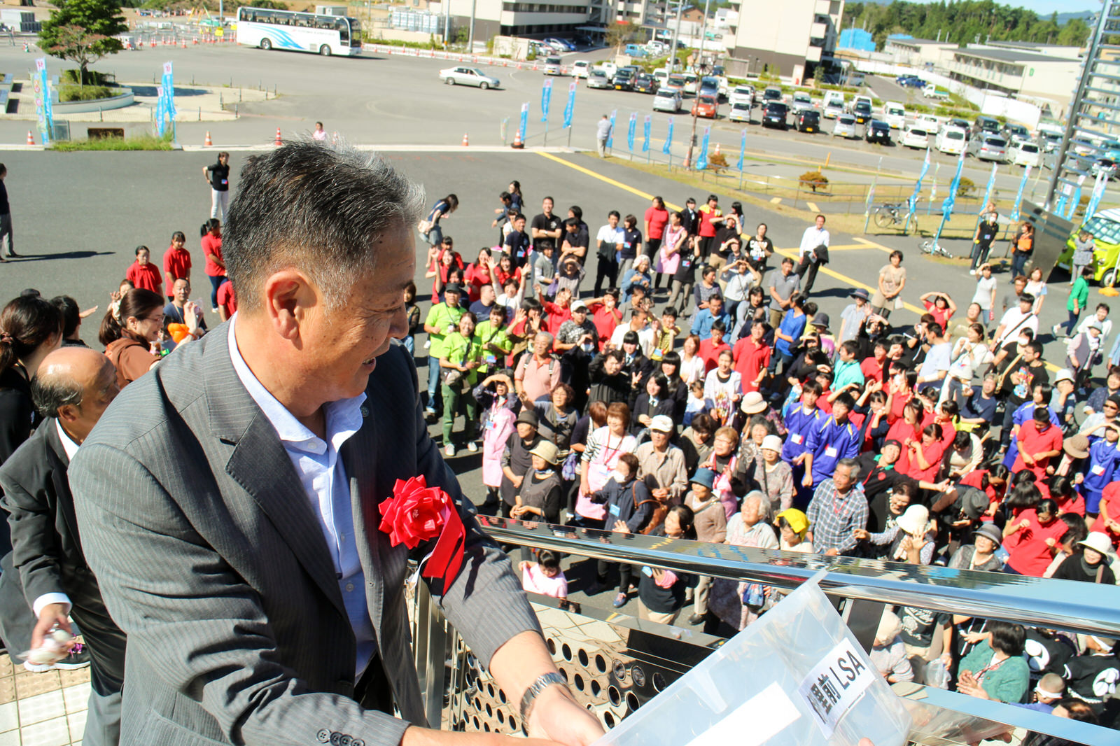 階段の上からスーツ姿の副町長が笑顔で透明ケースの中の品物を手にし、下に集まった大勢の観客に向かって配ろうとしている場面を上から捉えた餅まき準備の写真