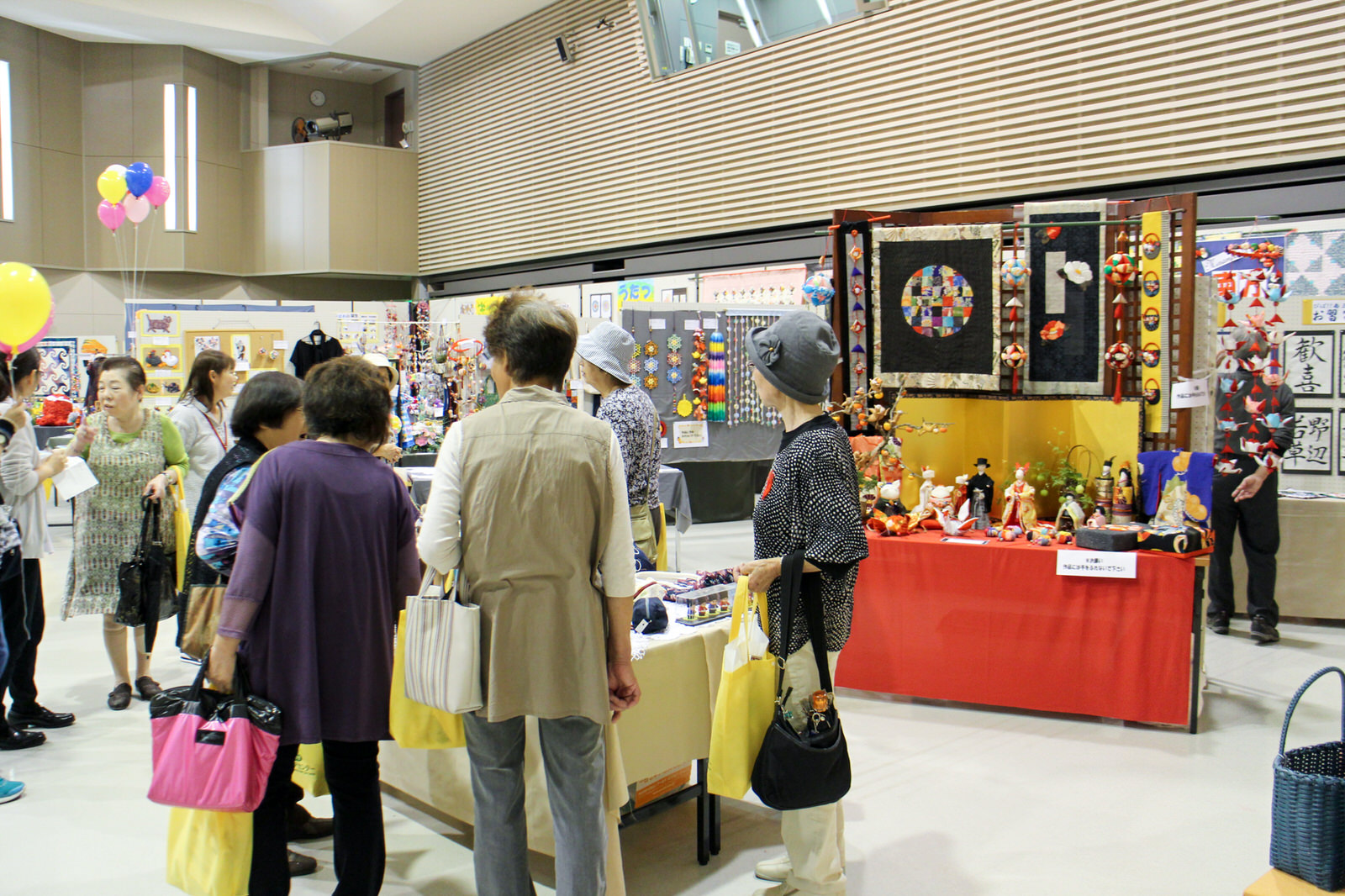 展示会場で手作りの和風の飾りや人形などが並ぶブースを複数の女性が興味深そうに見て回っている様子を捉えた、手工芸品展示コーナーの写真