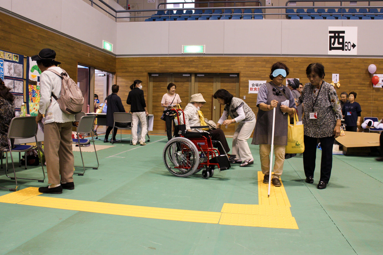 スタッフがサポートするなかいすの疑似体験をする女性と、体育館の通路に設置された黄色の点字ブロックの上を、目隠しをして白杖を使って歩く高齢の女性が、女性スタッフと共に歩行体験をしているバリアフリー体験コーナーの写真