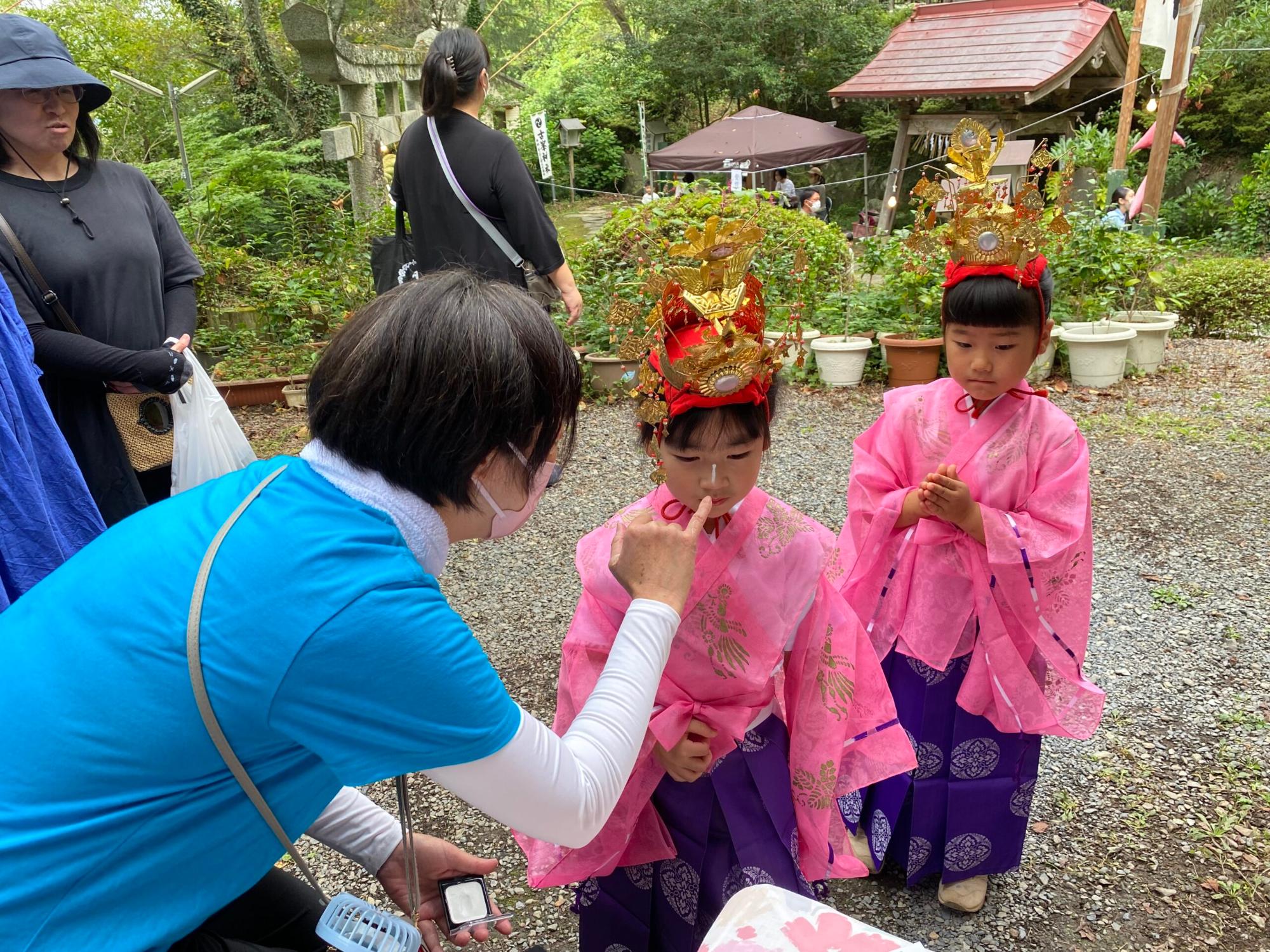 伝統衣装を着た女の子2人が並び、手前の大人が子どもの鼻筋に白い粉を塗っている様子の写真