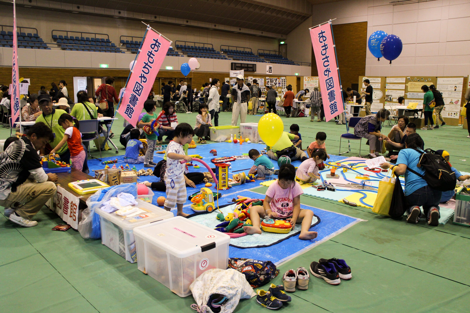 体育館の一角にブルーシートを敷き、大人たちが周囲で見守る中、たくさんの子どもたちがカラフルなおもちゃで遊び、ピンク色ののぼり旗に「おもちゃ図書館」と書かれているプレイスペースの様子を撮影した子ども向け遊びコーナーの写真