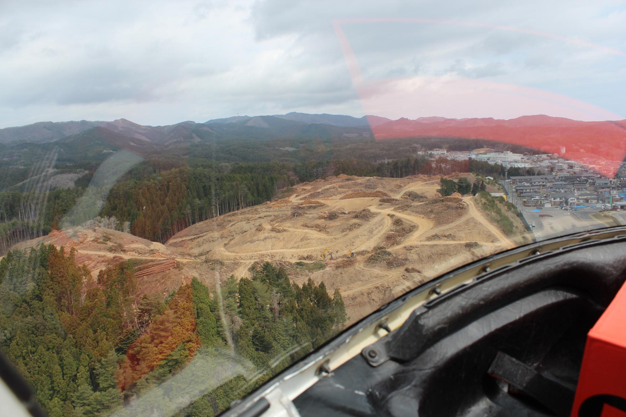 手前に伐採された木々と造成中の曲がりくねった道が広がり、奥には住宅地と山並みが見えるヘリコプターの中から撮影された写真