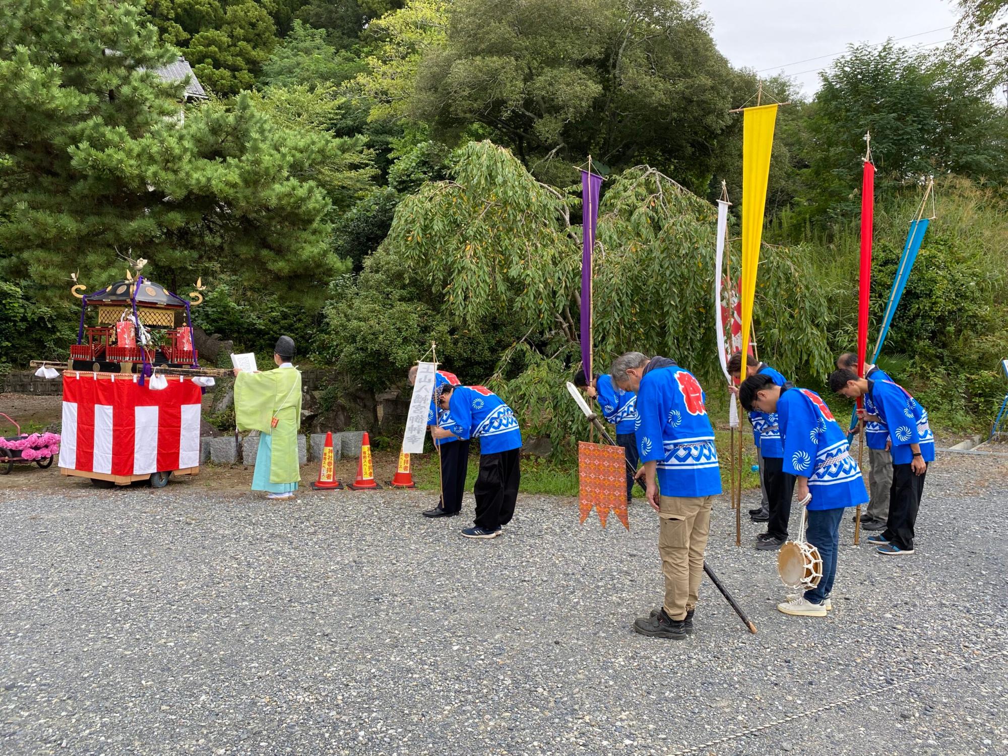 神職の男性が前に立ち、青い法被を着た数名の男性が深くお辞儀をしている、屋外での祭礼の写真