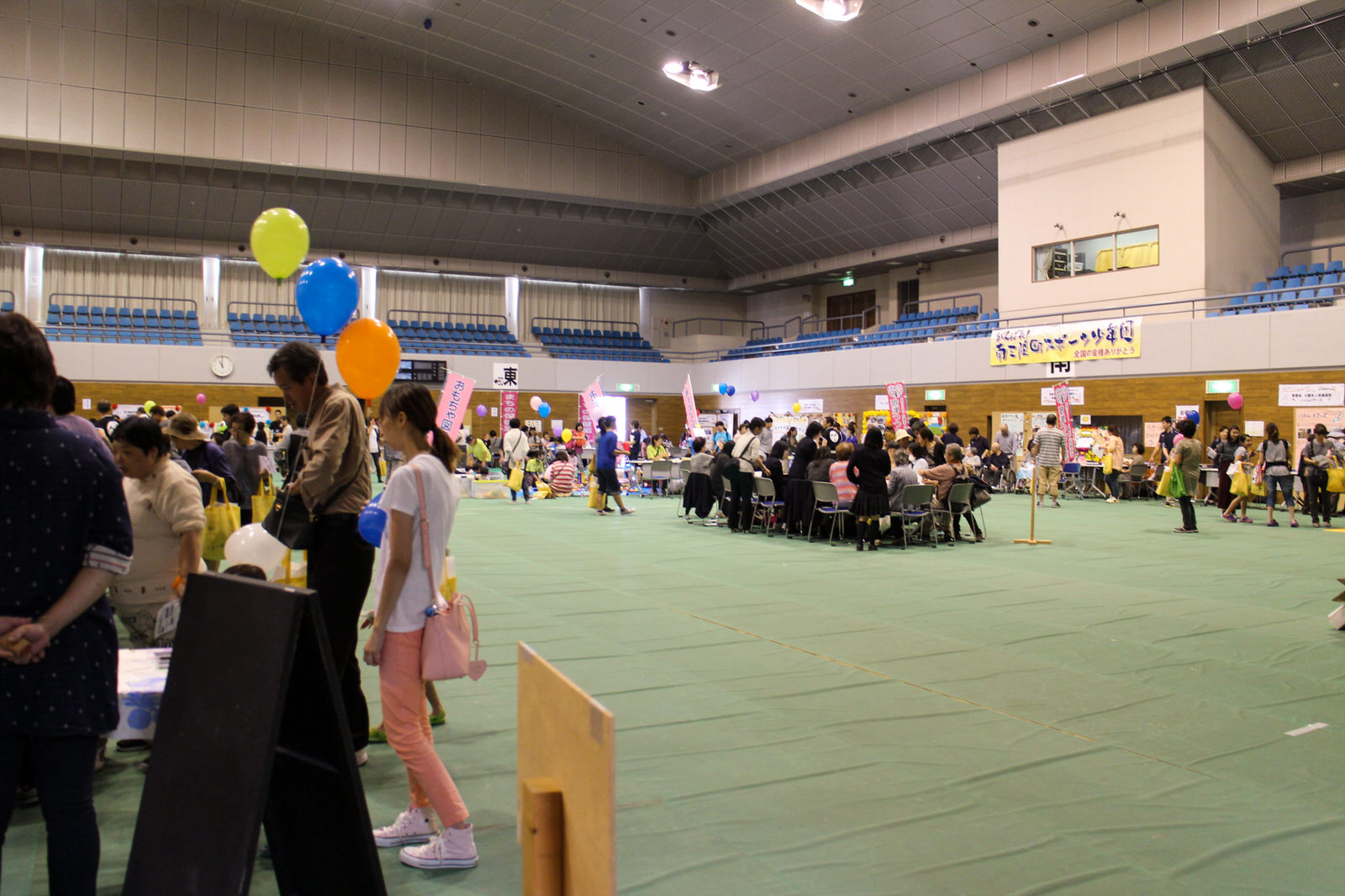 体育館のような広い屋内会場に色とりどりの風船が飾られた中、来場者が各ブースを訪れてイベントを楽しんでいる様子の写真
