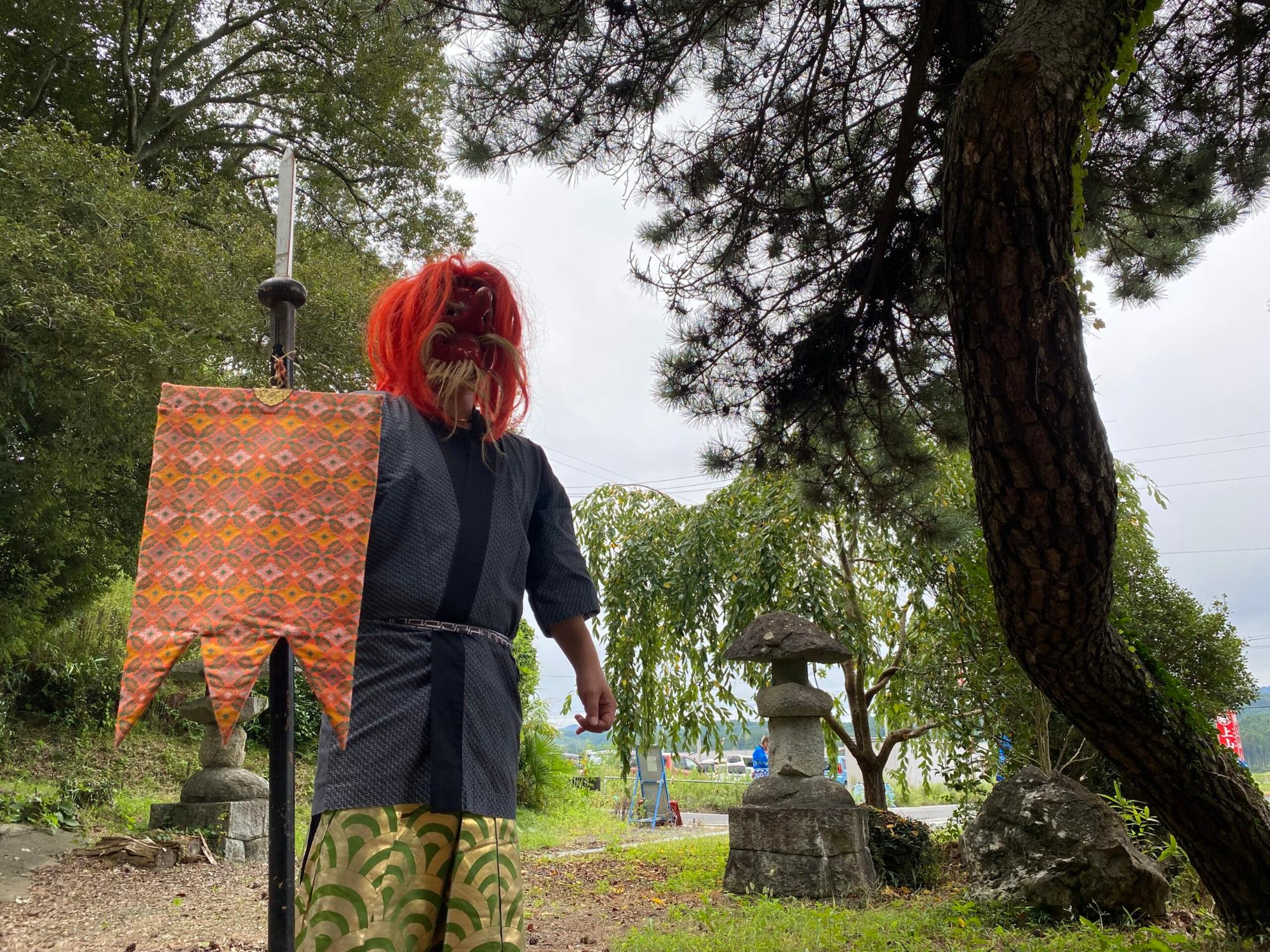 赤い髪と面をつけた伝統的な天狗の装束の男性が旗を持ち、木陰の下に立っている写真