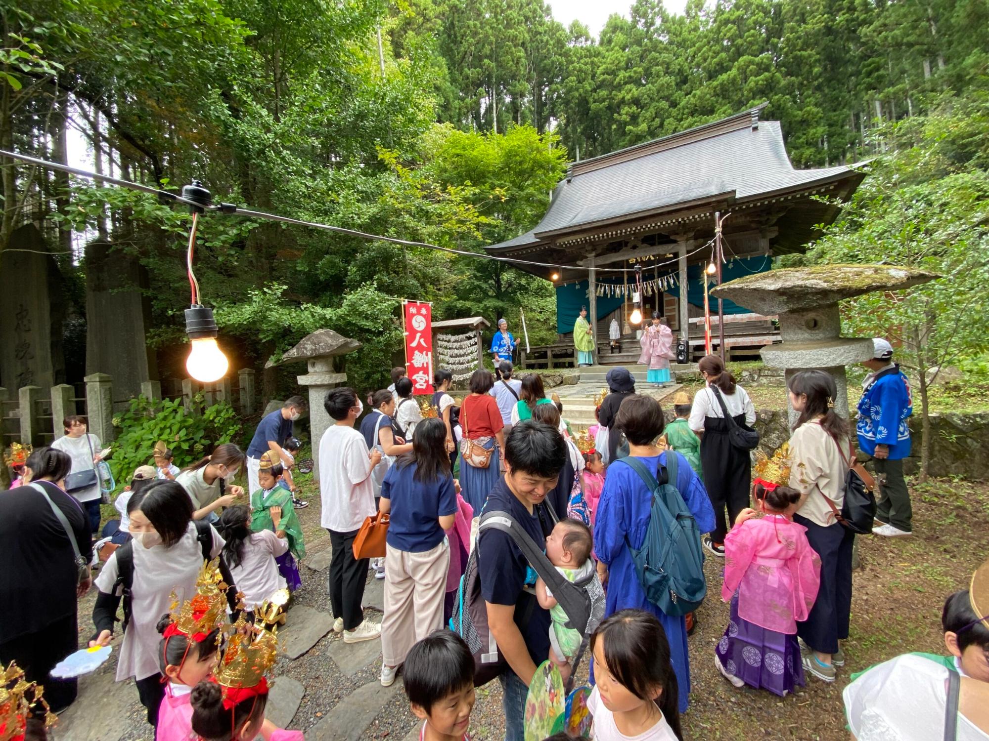 森の中の神社の前に子どもたちも伝統的な衣装で並び、大勢の人々が集まっている写真