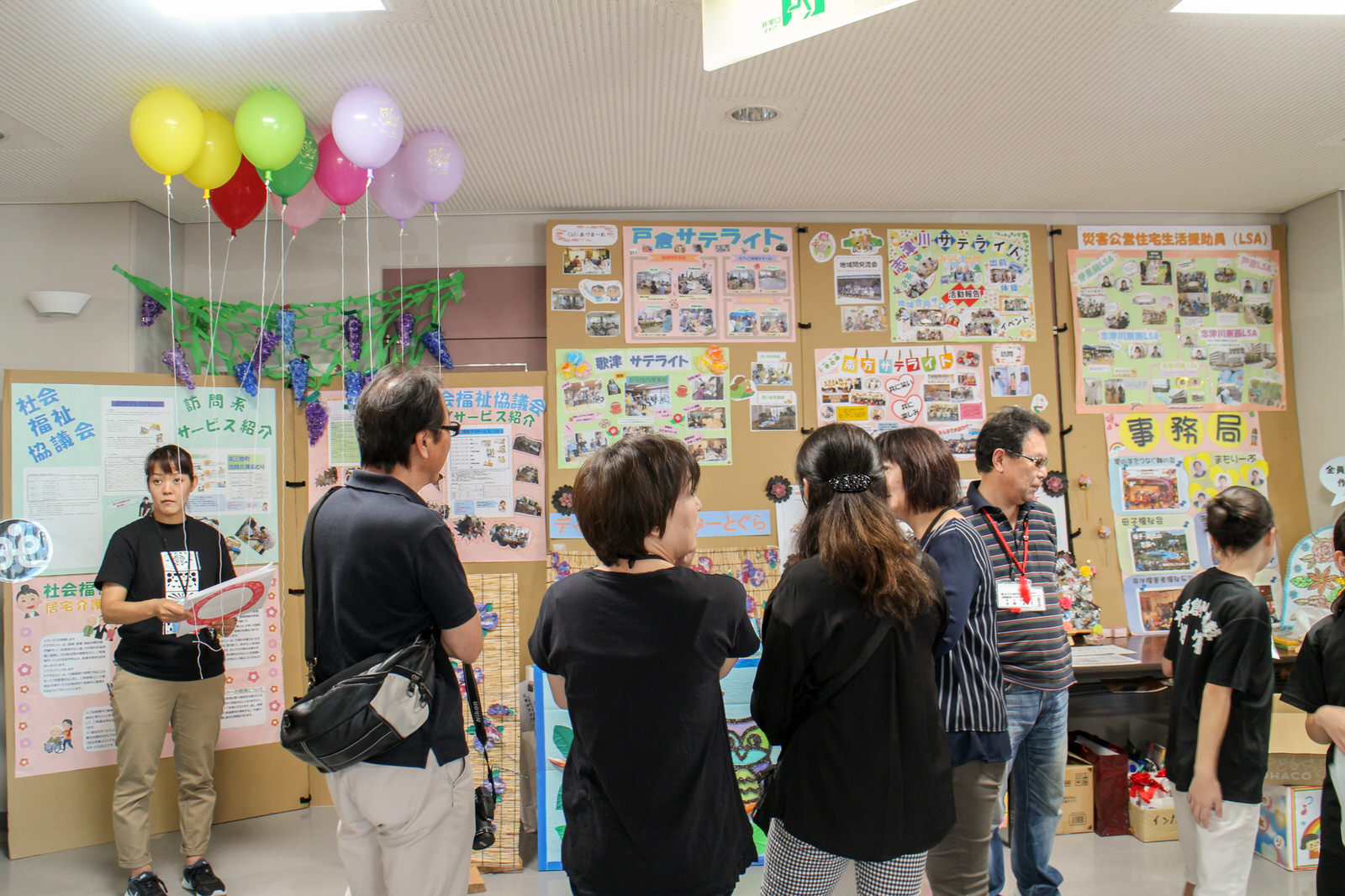 壁一面に貼られた活動紹介のポスターや掲示物の前で、説明を聞いたり交流したりしている来場者たちの写真