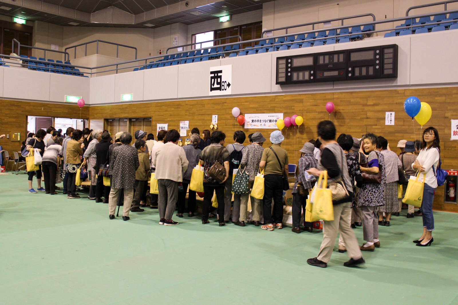 体育館のバザーを行っているブースで、黄色い手提げ袋を持った参加者たちが集まり品定めをしている様子を写した写真