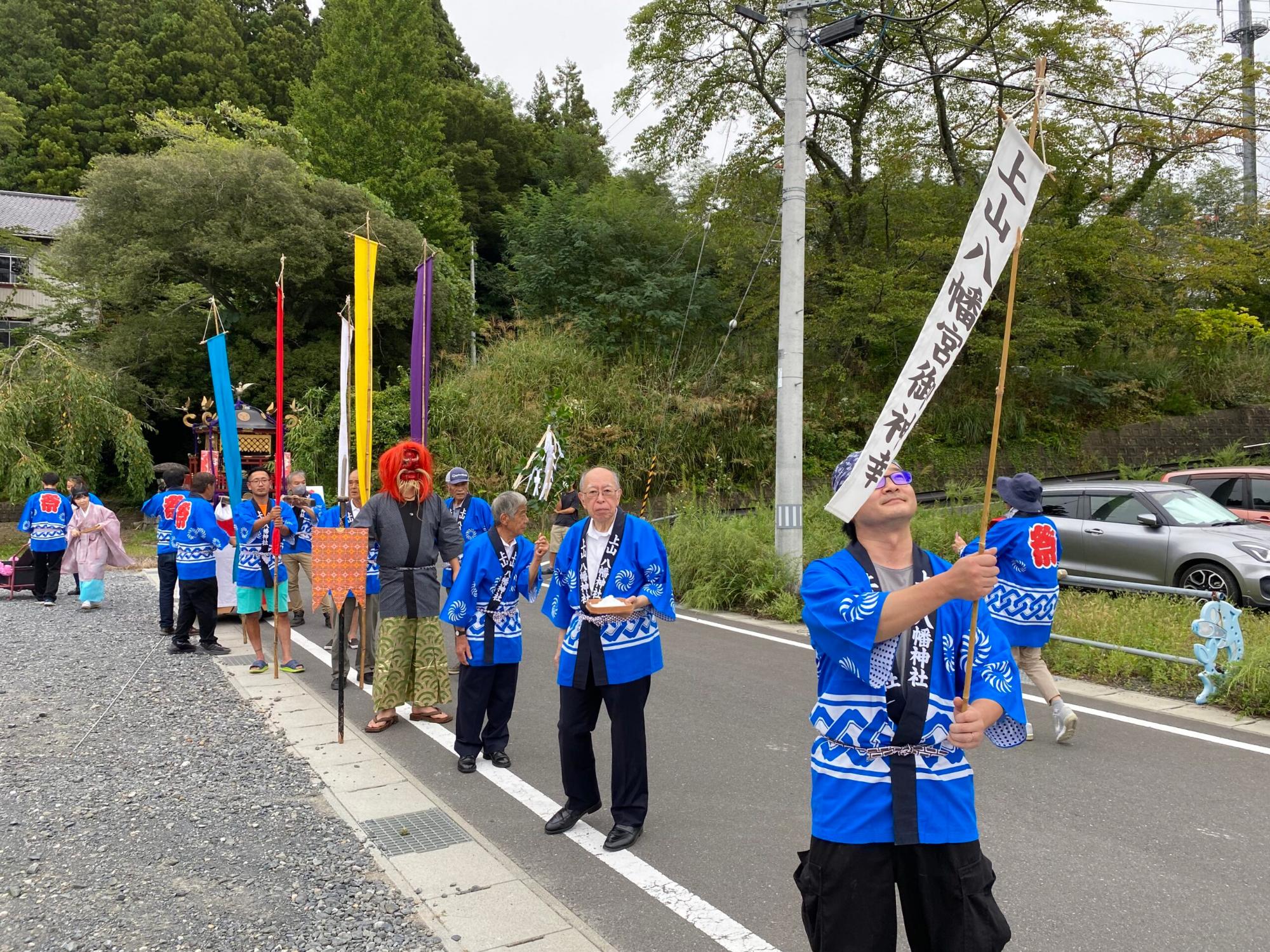 中央には赤い面をつけた人がおり、青い法被を着た大人たちが旗を持ち、一列に並んで歩いている写真