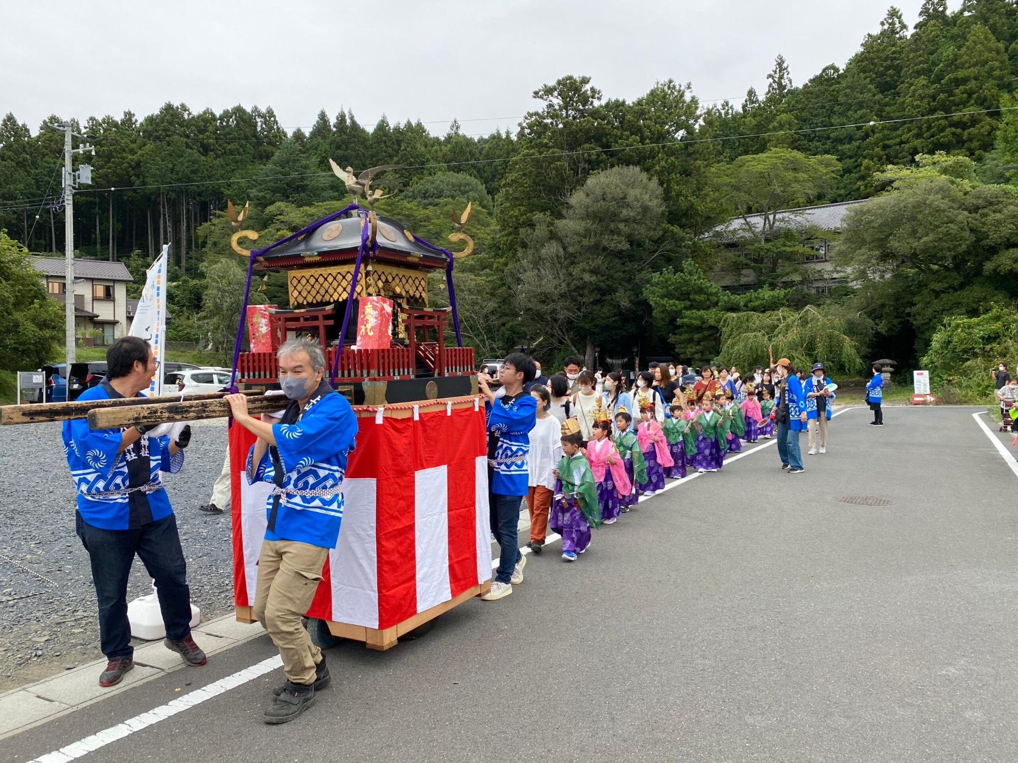 数人の大人が赤と白の布で飾られた神輿を担いでいる後ろに、色とりどりの衣装を着た子どもたちが並んでいる写真