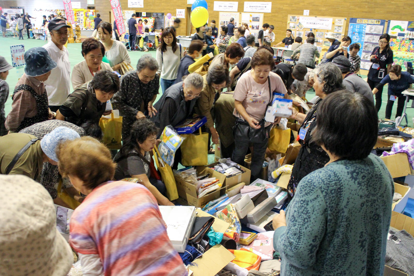 室内で多くの高齢者たちが段ボールに入った衣類や日用品などを熱心に選びながら手に取っている様子を捉えた、福祉バザーの買い物風景の写真
