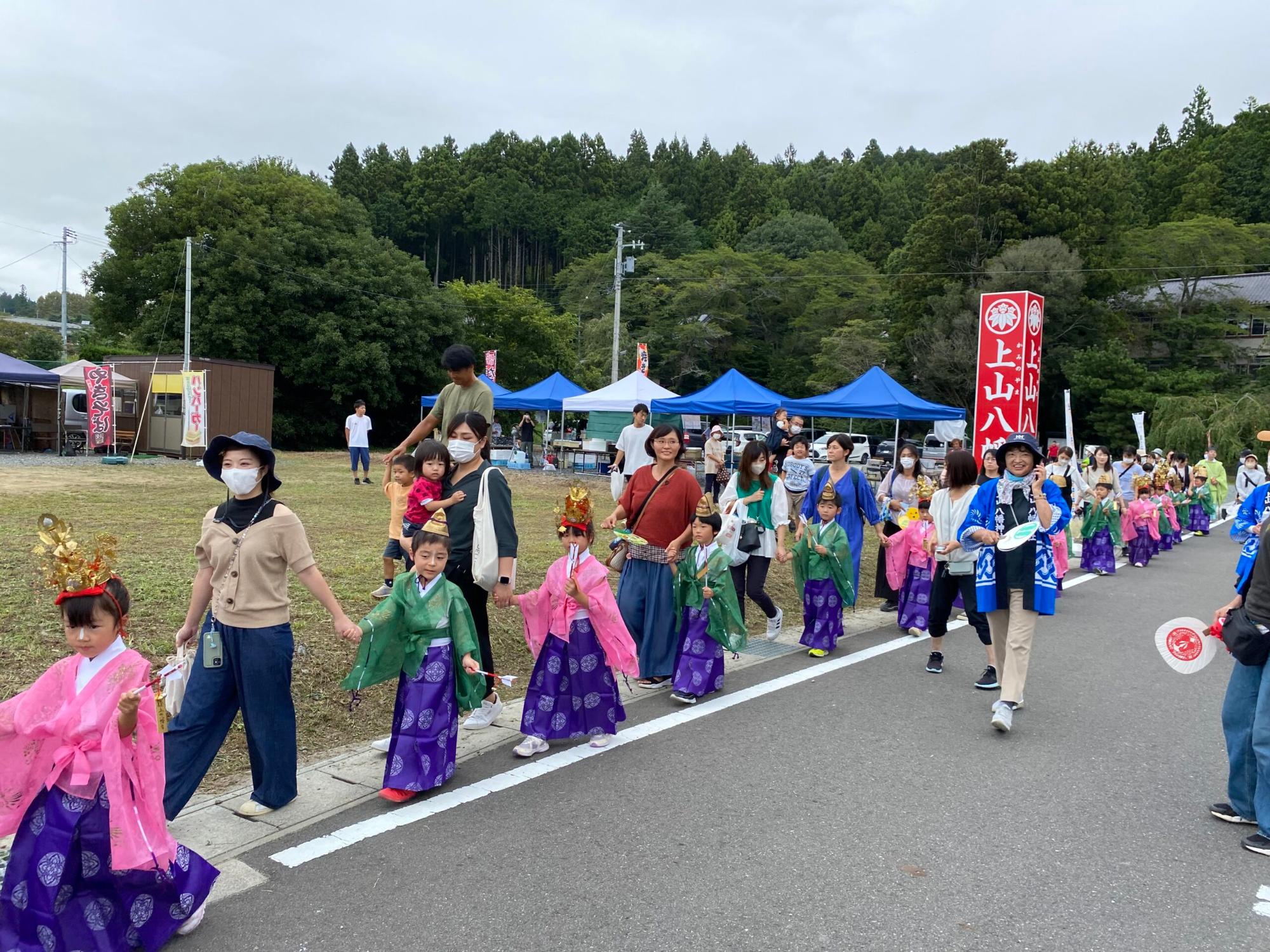 背景にはテントが並び、子どもたちが伝統的な和服を着て、大人と手をつなぎながら歩いている様子の写真