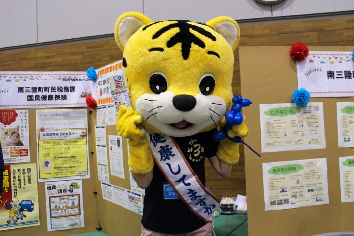 展示パネルなどが並ぶ室内の福祉イベント会場で、首に「健康してますか？」と書かれたたすきをかけた大きな黄色い虎のキャラクターが、片手にバルーンアートを持ち立っている写真