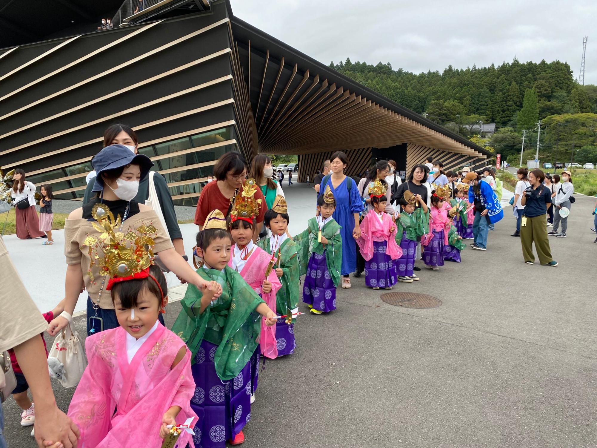 子どもたちが伝統的な衣装を身にまとい、大人と手をつないで行列を作って歩いている様子の写真