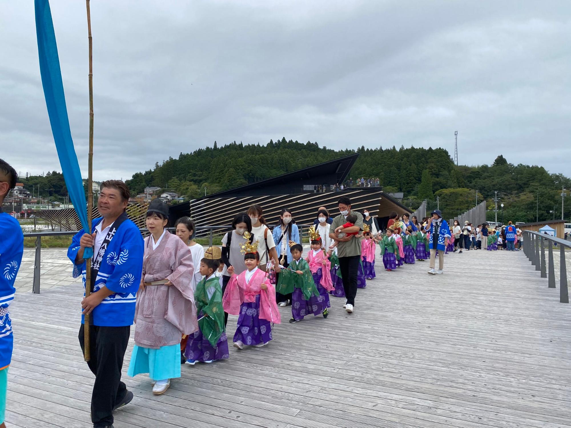 法被を着た男性を先頭に、子どもたちが伝統的な衣装を身にまとい、大人と手をつないで行列を作って歩いている様子の写真