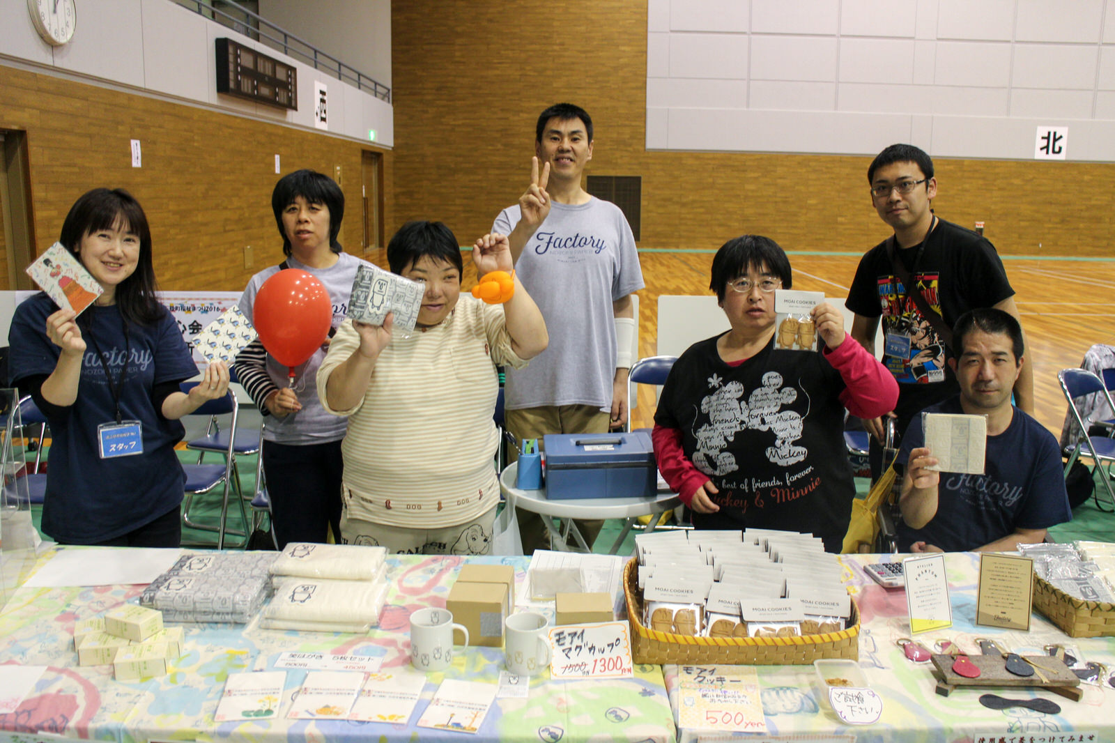 テーブルに並べられた手作り雑貨やお菓子を前に、7人の男女が笑顔で商品を手に取りながらアピールしている、障害のある人々が運営する福祉作業所の販売ブースの写真