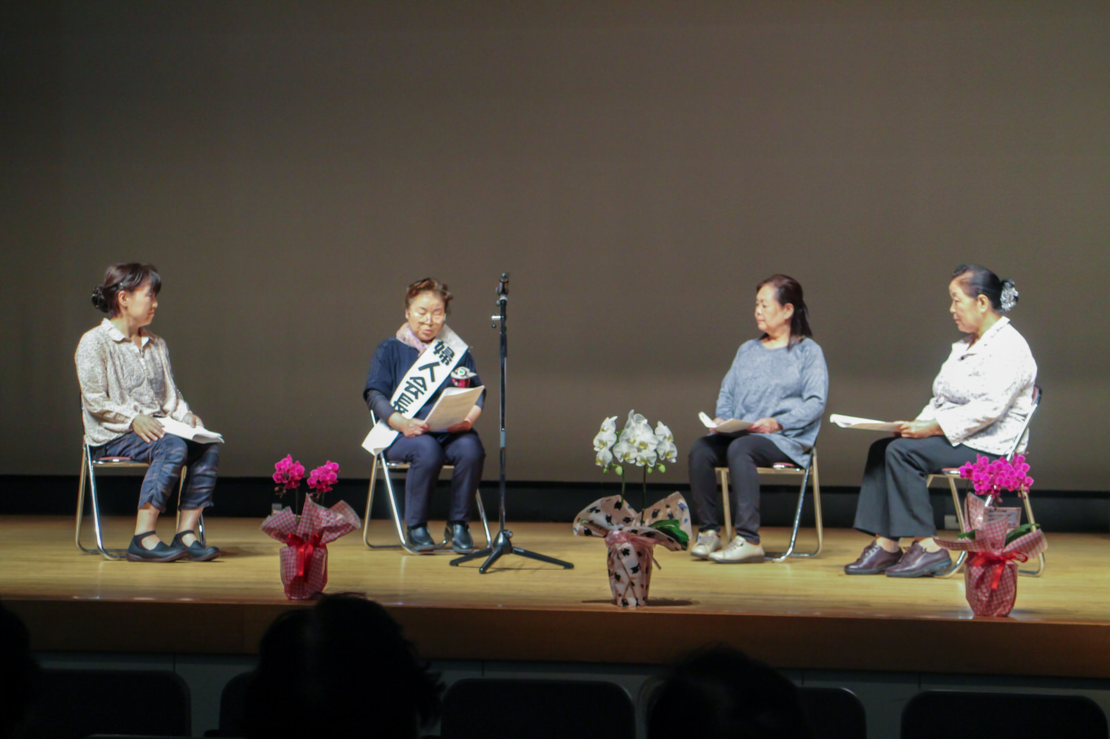 ピンクと白色の鉢植えの花が並んでいる舞台の上で、中央のマイクを囲んで並べた椅子に4人の女性が座り台本を読みながら寸劇を行っている様子の写真